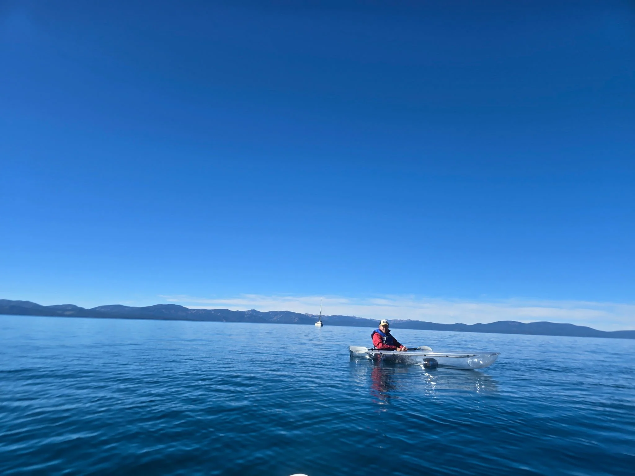 A person in a small boat on a large body of water with a sailboat anchored in the distance, and mountains on the horizon under a clear blue sky.