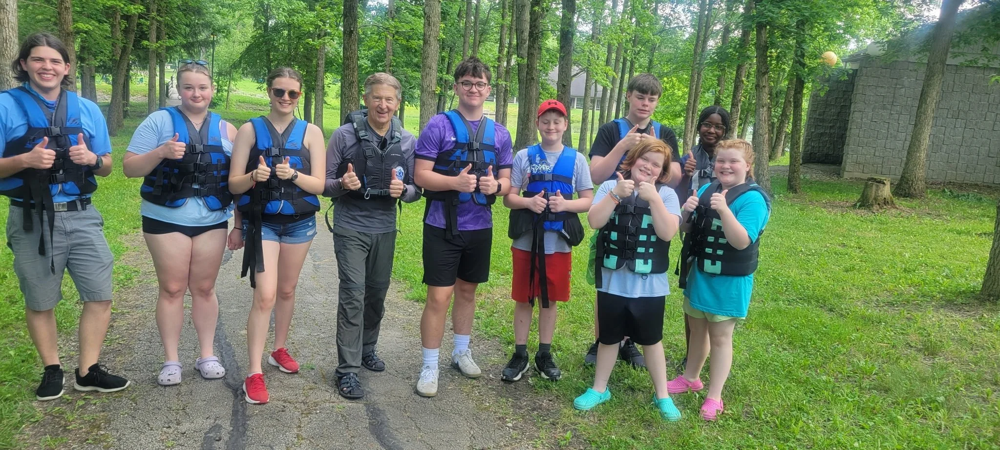 Group of children and an adult smiling and giving thumbs-up, standing outdoors on a trail in a wooded area, all wearing life jackets, indicating they are on a raft trip or outdoor adventure.
