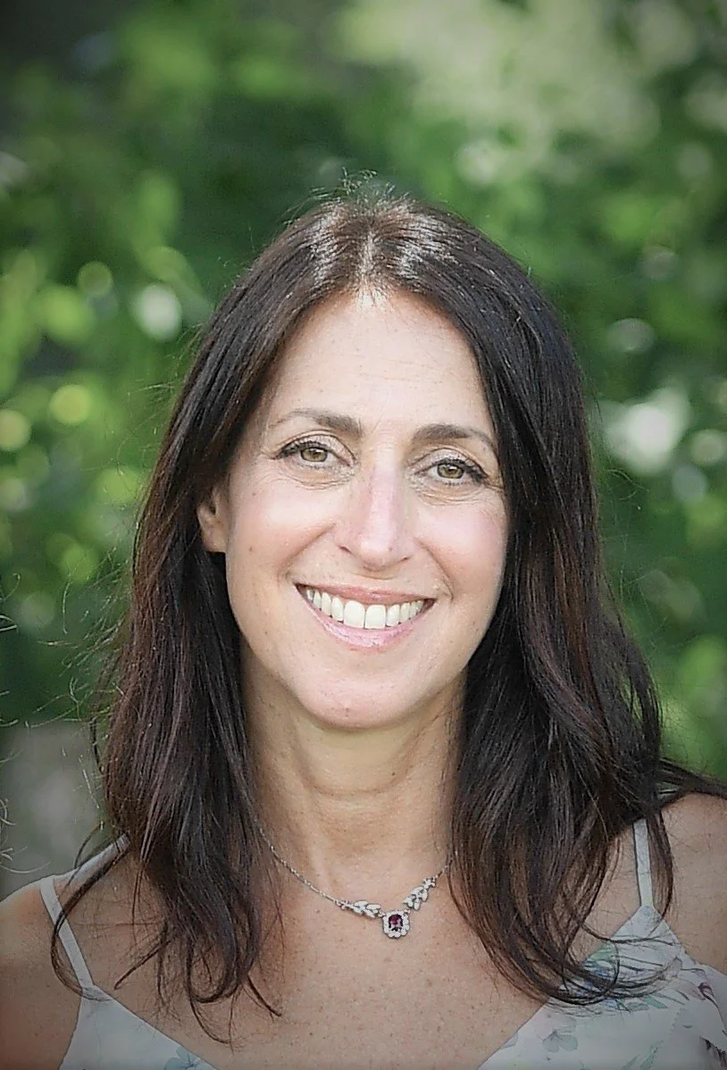A woman with shoulder-length dark brown hair smiling outdoors with a blurred green background.