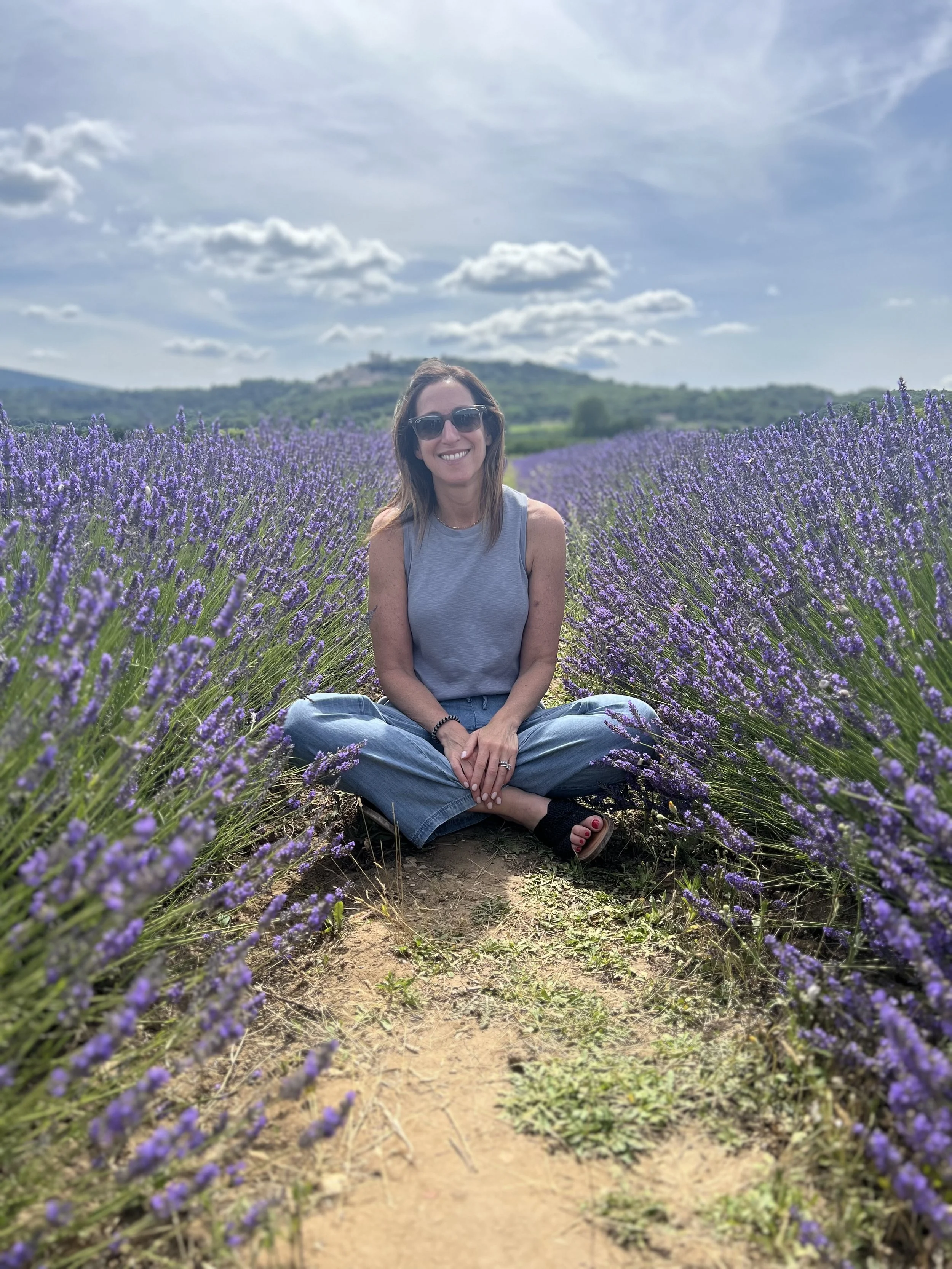 A woman sitting cross-legged in a field of blooming lavender flowers, wearing sunglasses and a sleeveless top, smiling at the camera on a sunny day with a partly cloudy sky and hills in the background.