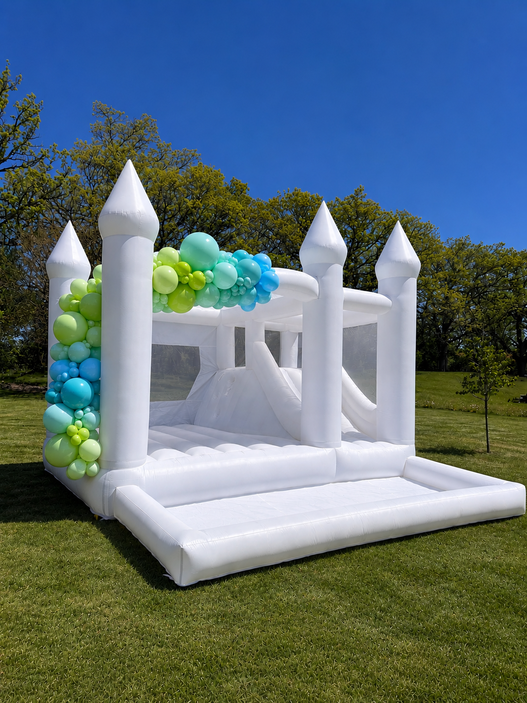 White inflatable castle with four towers, decorated with green and blue balloons, set up outdoors on a grassy field with trees and blue sky in the background.