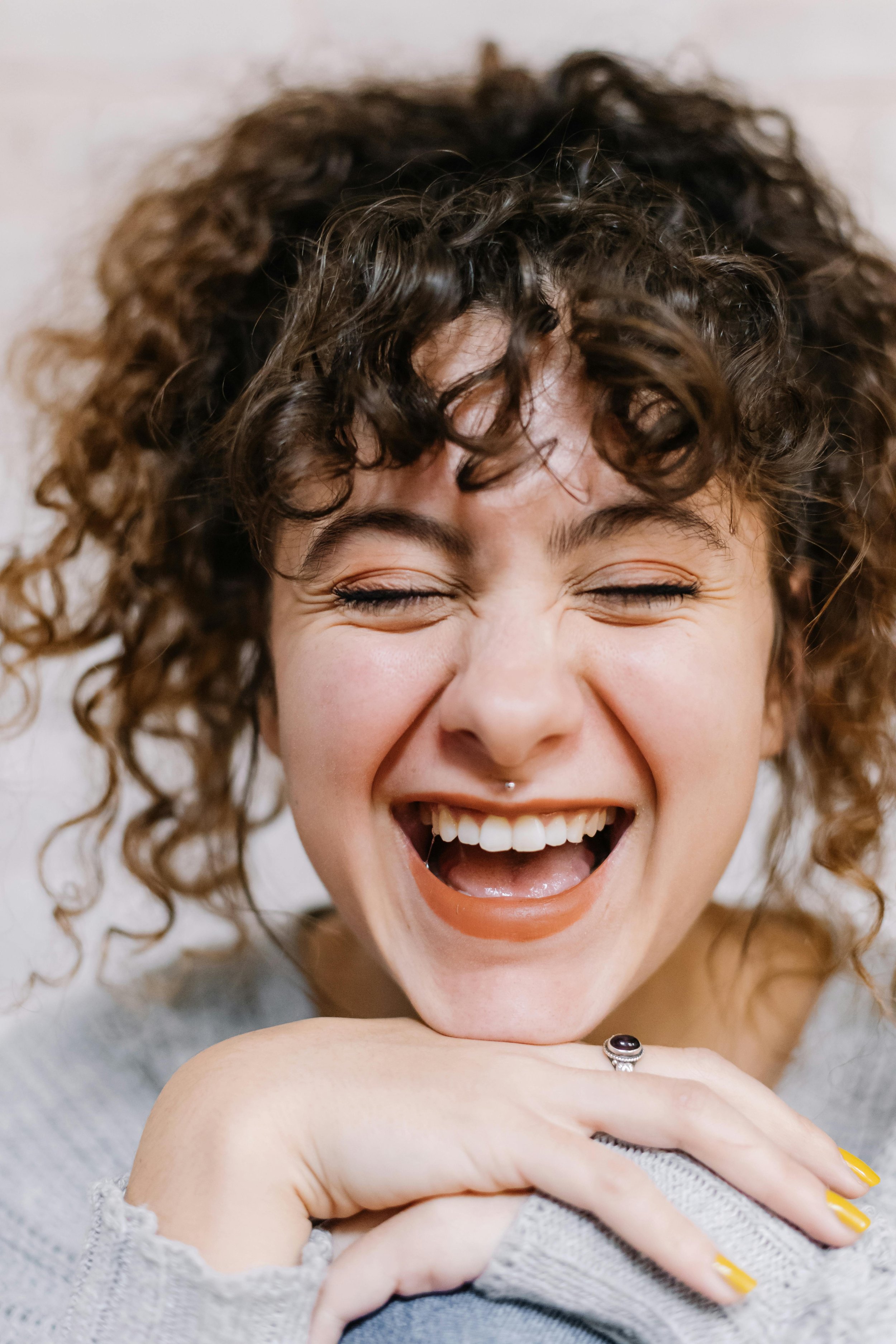 Person with curly hair laughing joyfully, head tilted back, eyes closed, wearing a ring, and a gray knitted top.