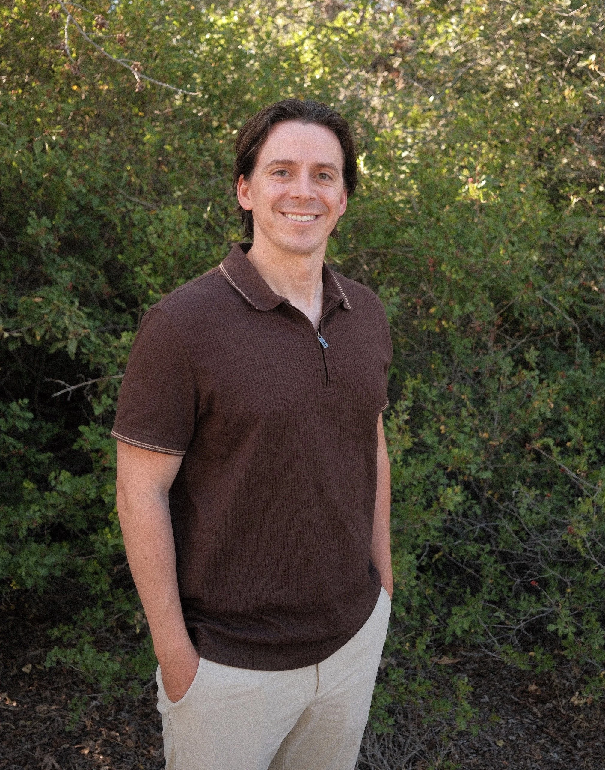 A man standing outdoors in front of green foliage, wearing a brown zip-up shirt and beige pants, smiling at the camera.