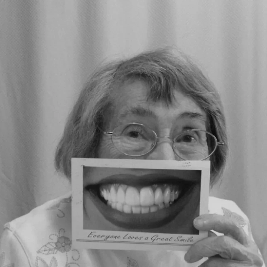 A woman with glasses holding a photo of a big smiling mouth in front of her face, with the caption "Everyone Loves a Great Smile."