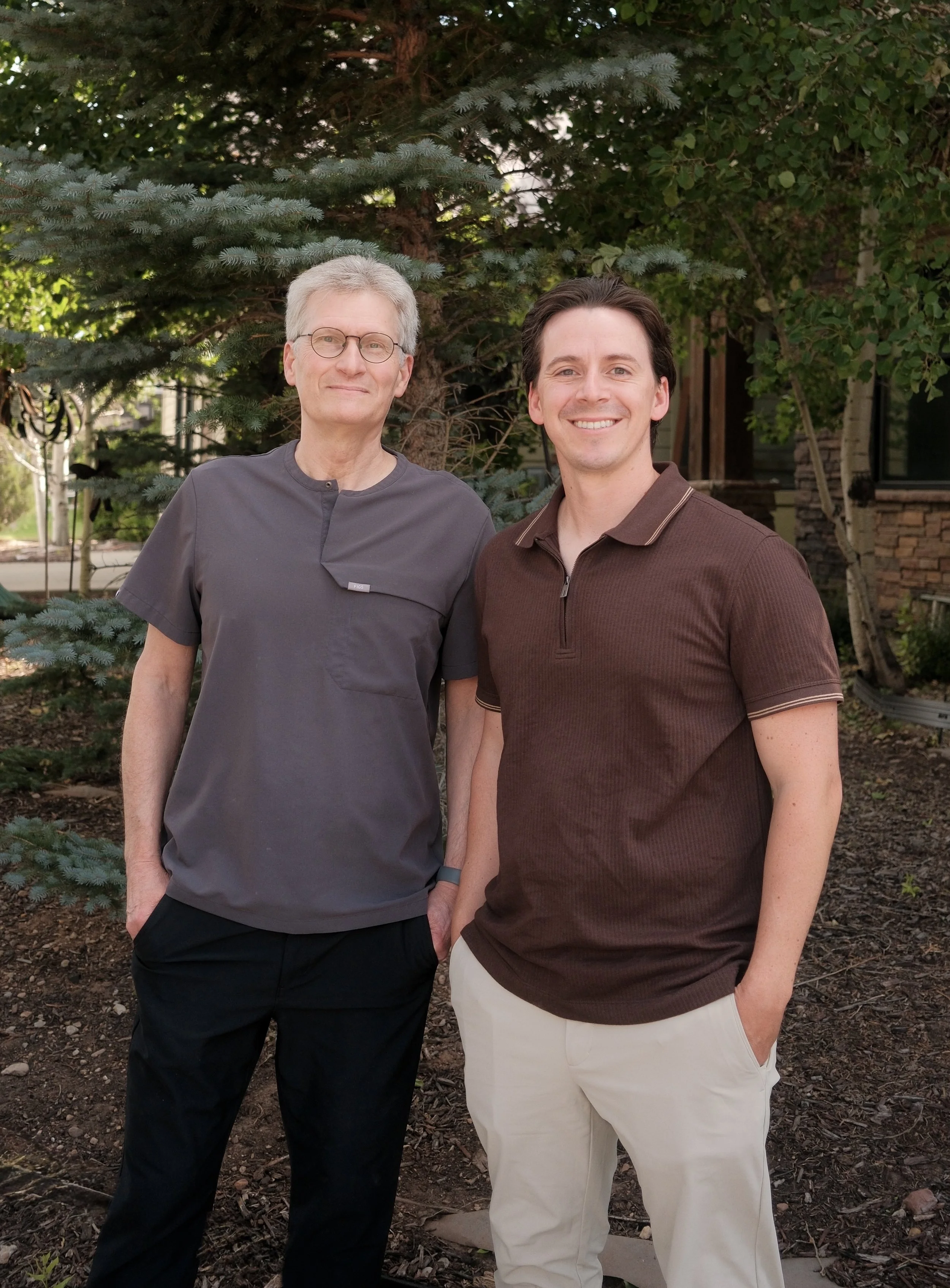 Two men standing outdoors in front of greenery, smiling. The man on the left is wearing glasses, a gray medical-style scrub top, and black pants. The man on the right has dark hair, a brown polo shirt, and beige pants. They are standing close together with their hands resting in their pockets, in a garden or yard setting.