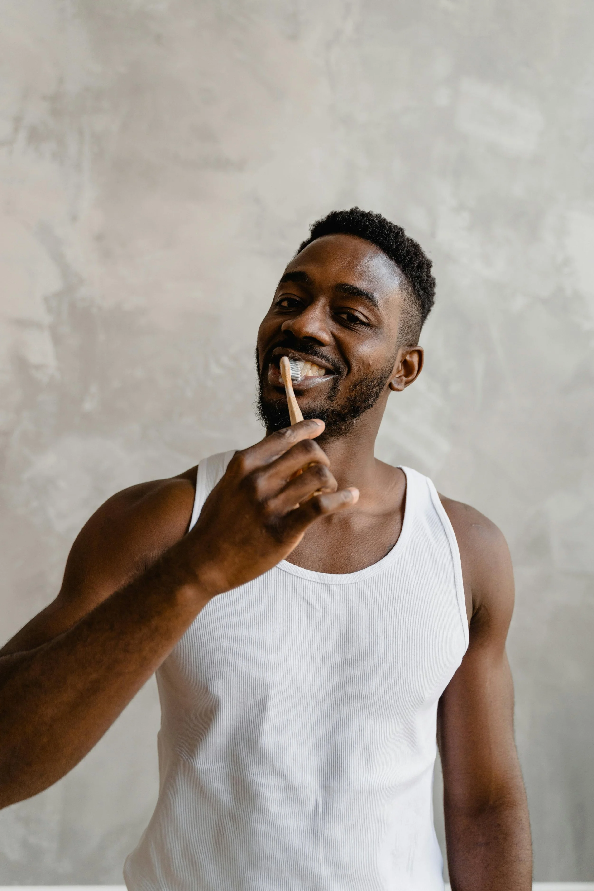 A man with dark skin wearing a white tank top brushing his teeth with a toothbrush.