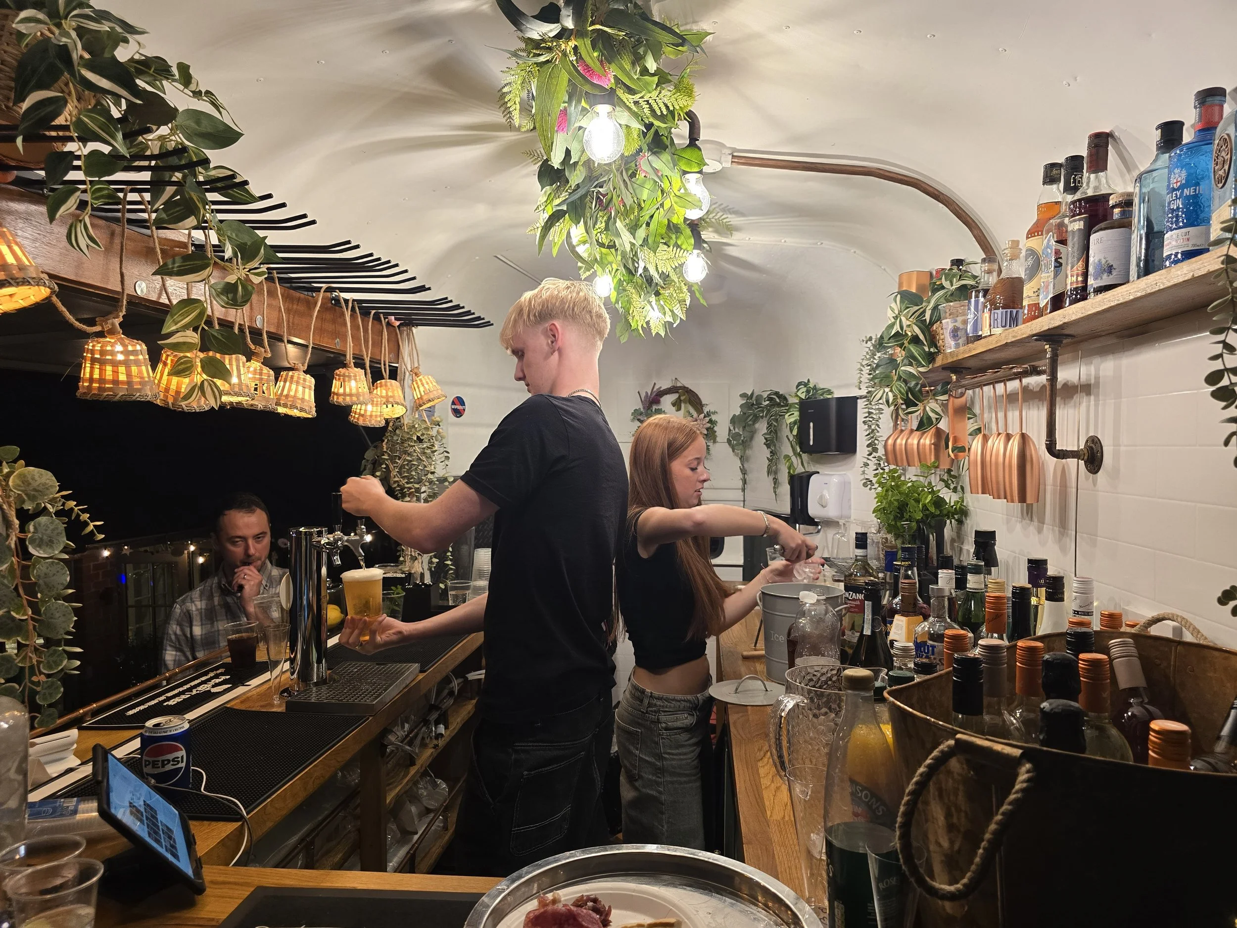 People working behind a bar with liquor bottles and bar tools, decorated with hanging plants and warm lighting.
