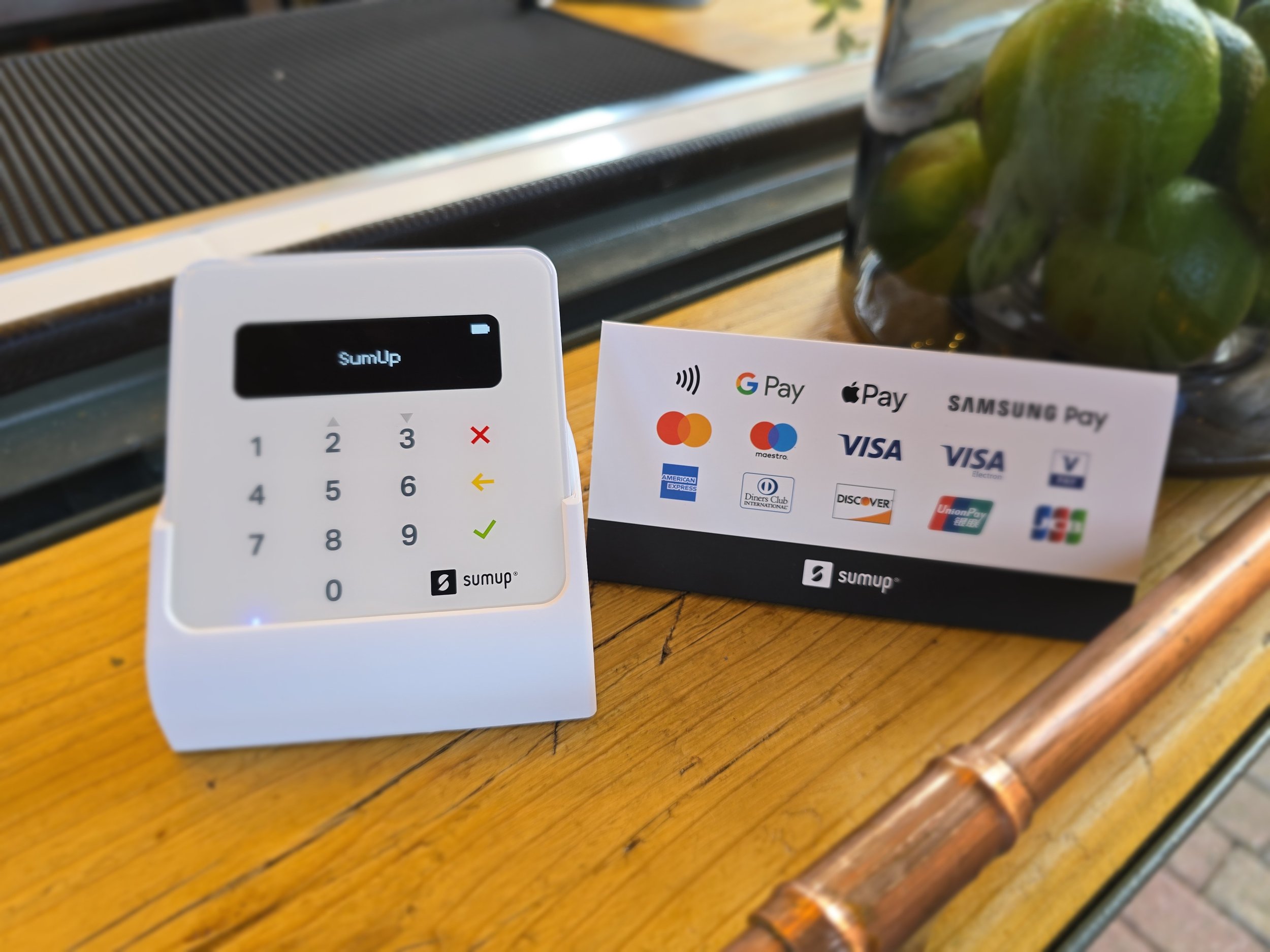 A small white electronic device with a keypad displaying the words 'SunUp' and a card with various payment logos including Google Pay, Apple Pay, Mastercard, Visa, and others, placed on a wooden surface next to a black object and a glass container with green limes.