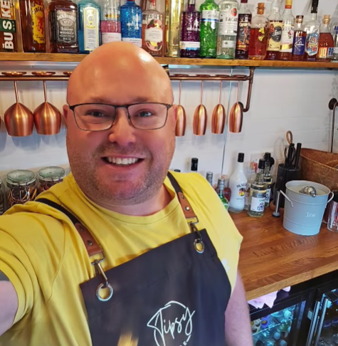A man smiling in a bar or kitchen area with a variety of liquor bottles on shelves behind him, wearing glasses, a yellow T-shirt, and a black apron with a logo.