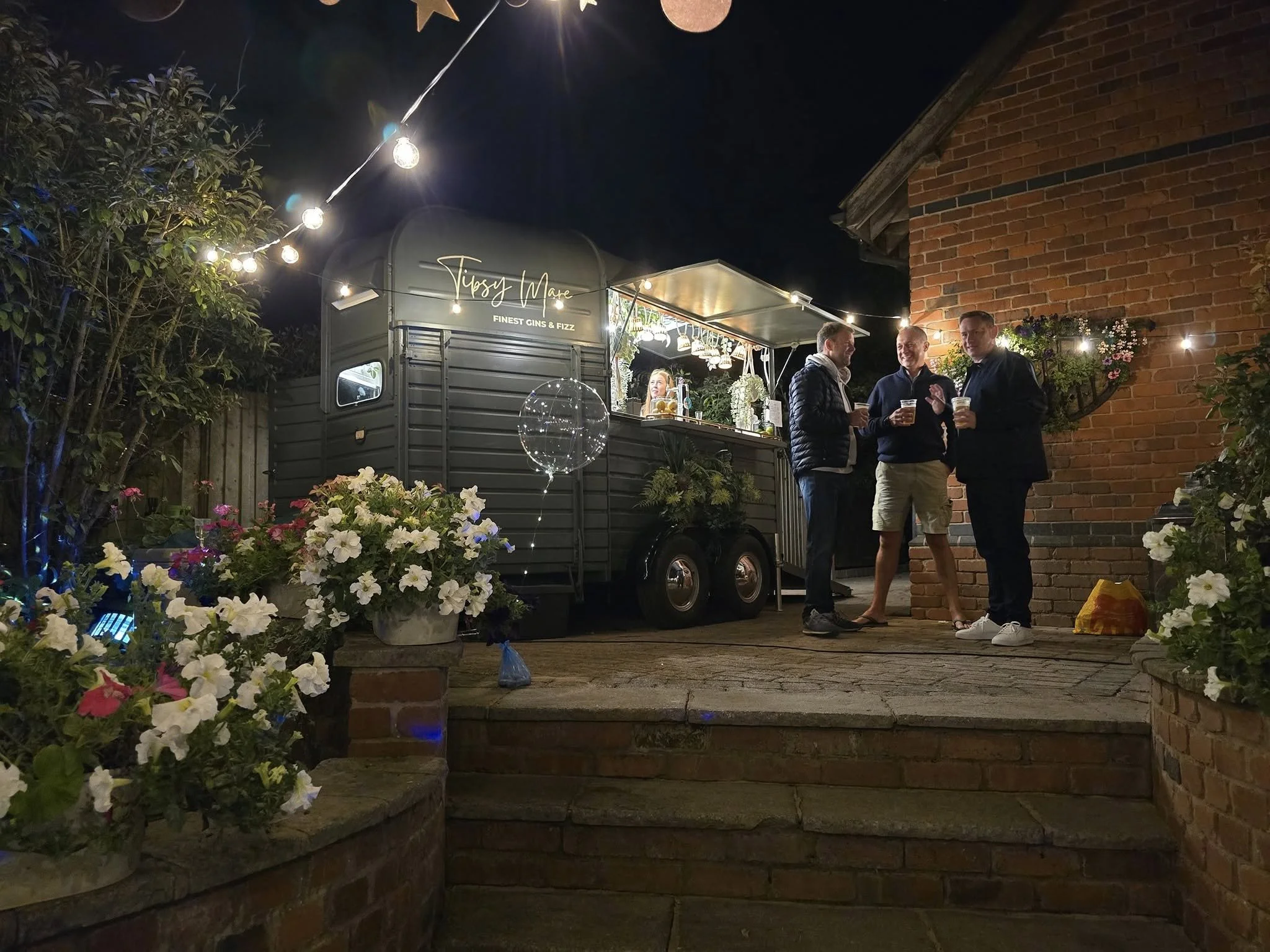 Night scene of an outdoor gathering with a mobile bar named 'Tipsy Made' serving drinks, illuminated by string lights, with three men holding drinks and chatting near a red brick wall. There are flowers and potted plants in the foreground.