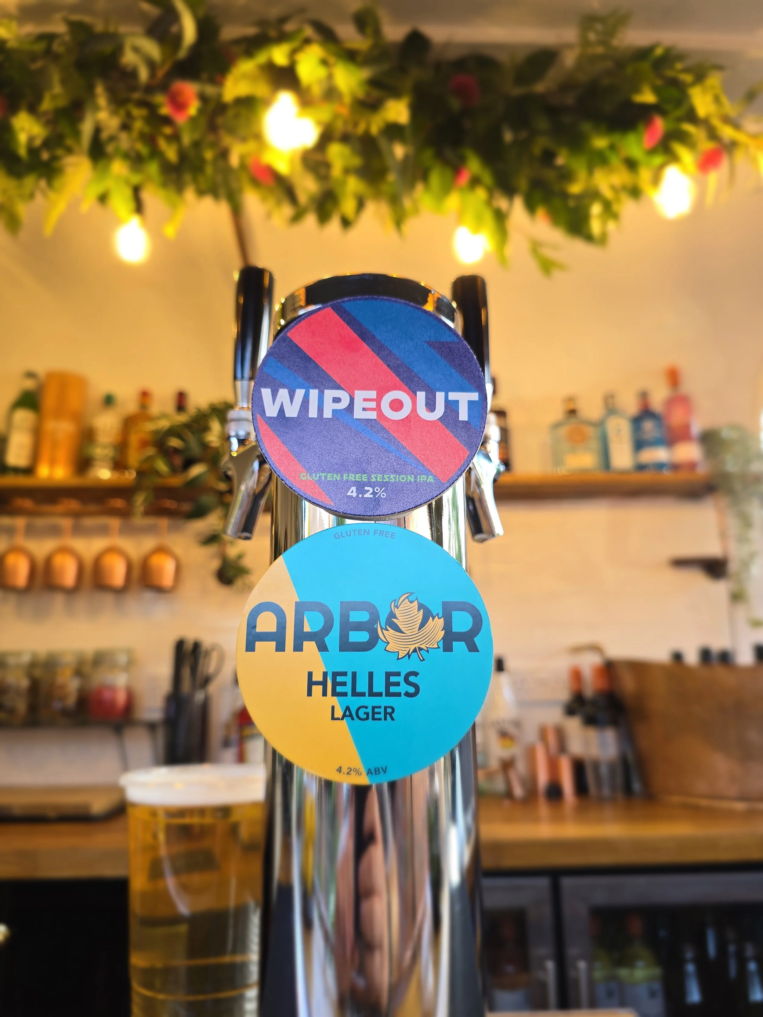 Close-up of beer tap handles at a bar, with two of the handles having round labels: one purple and pink labeled 'WIPEOUT' indicating a gluten-free session IPA with 4.2% alcohol content, and one blue and yellow labeled 'ARBOUR Helles Lager' with 4.2% ABV. The background features a bar with various bottles on shelves, and string lights hanging above.