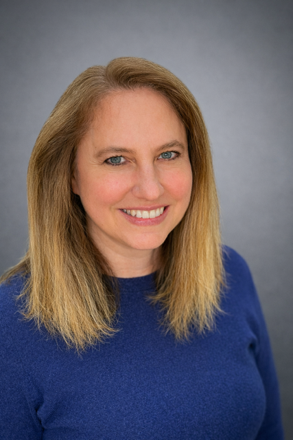 Close-up portrait of a smiling woman with blonde hair in a blue top against a gray background.