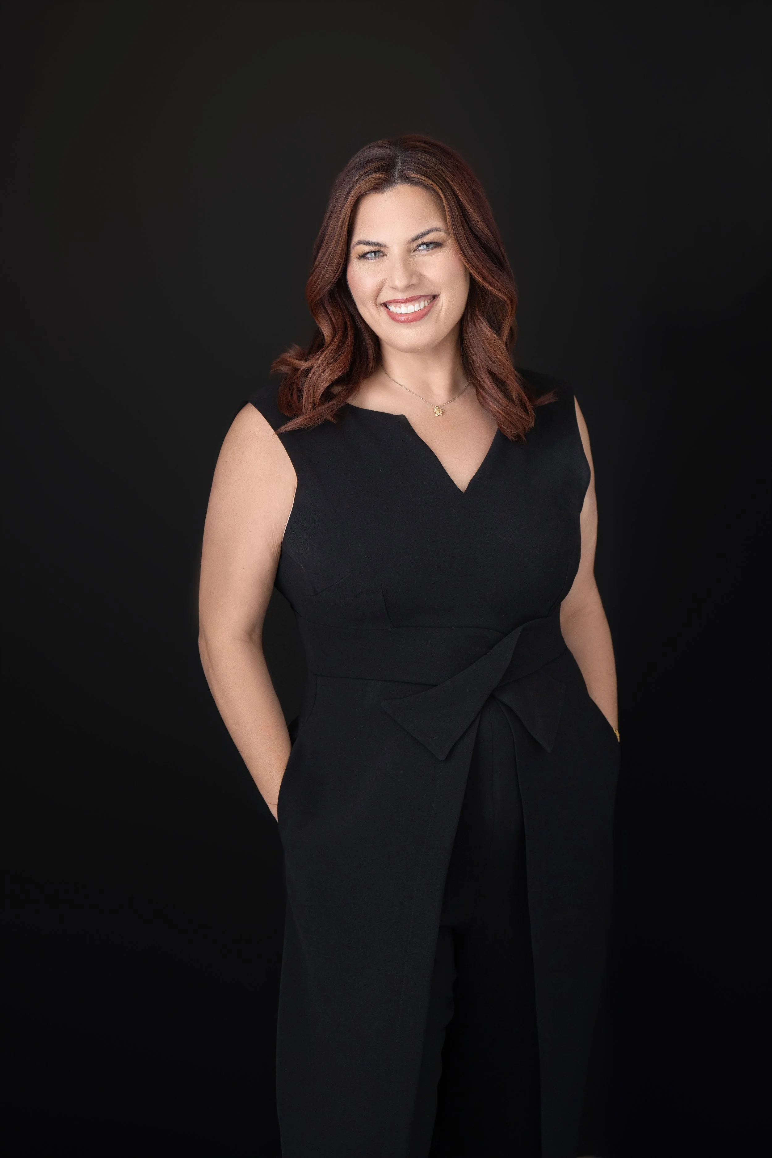 A woman with shoulder-length brown hair styled in loose waves, wearing a sleeveless black dress with a V-neckline, standing against a plain black background, smiling directly at the camera.