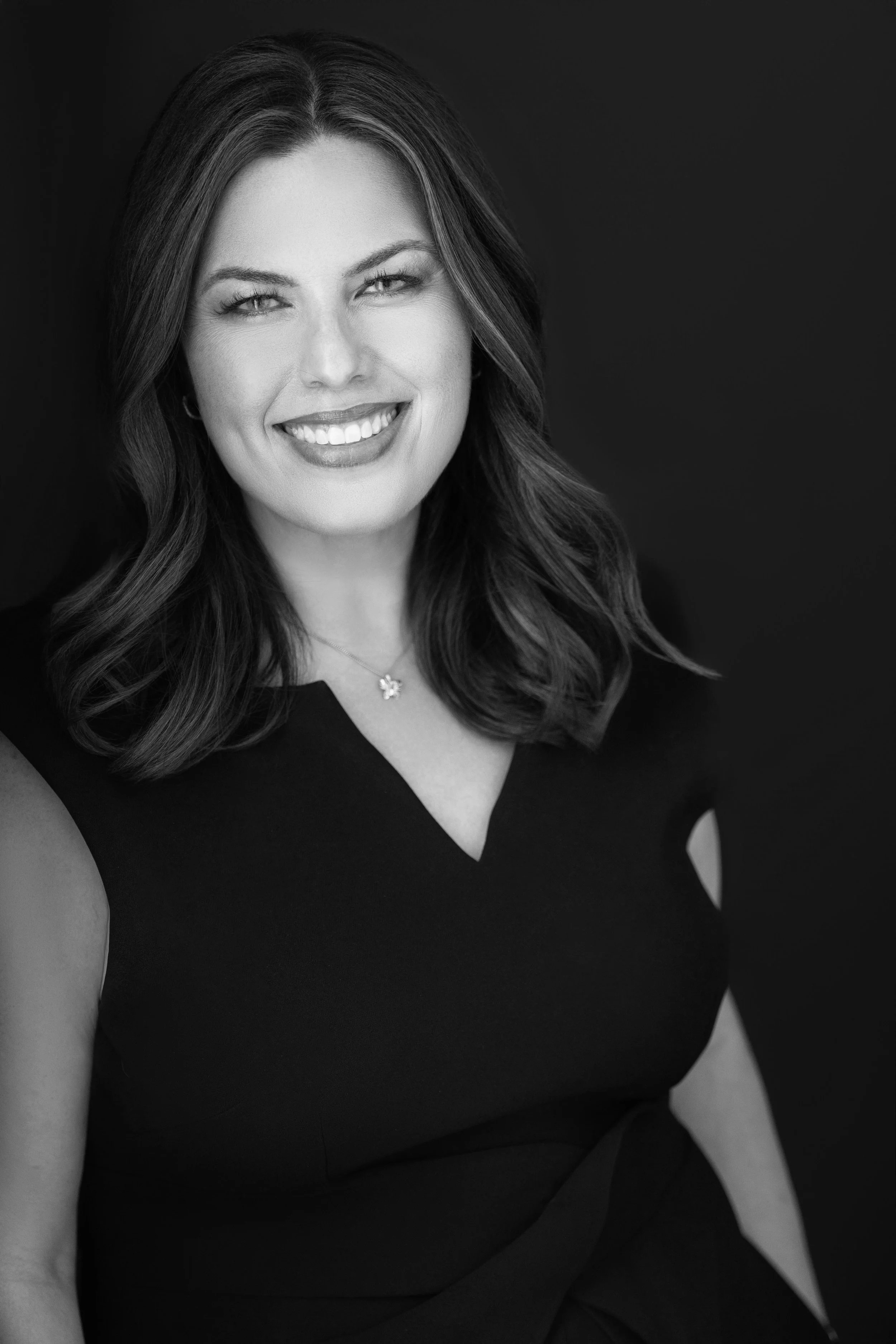 Black and white portrait of a woman with shoulder-length hair, smiling, wearing a sleeveless black top, a necklace with a butterfly pendant, and sitting against a dark background.