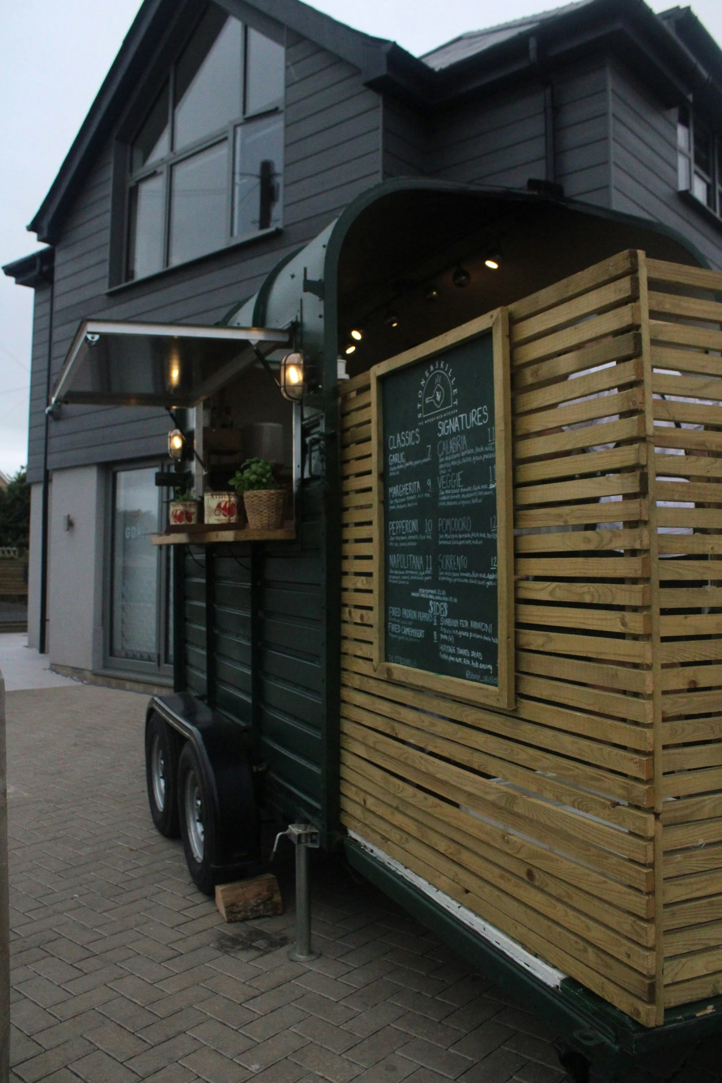 A food truck with a wooden lattice side panel, serving menu board, and small open window with potted plants and canned goods inside, parked on a brick sidewalk during dusk.