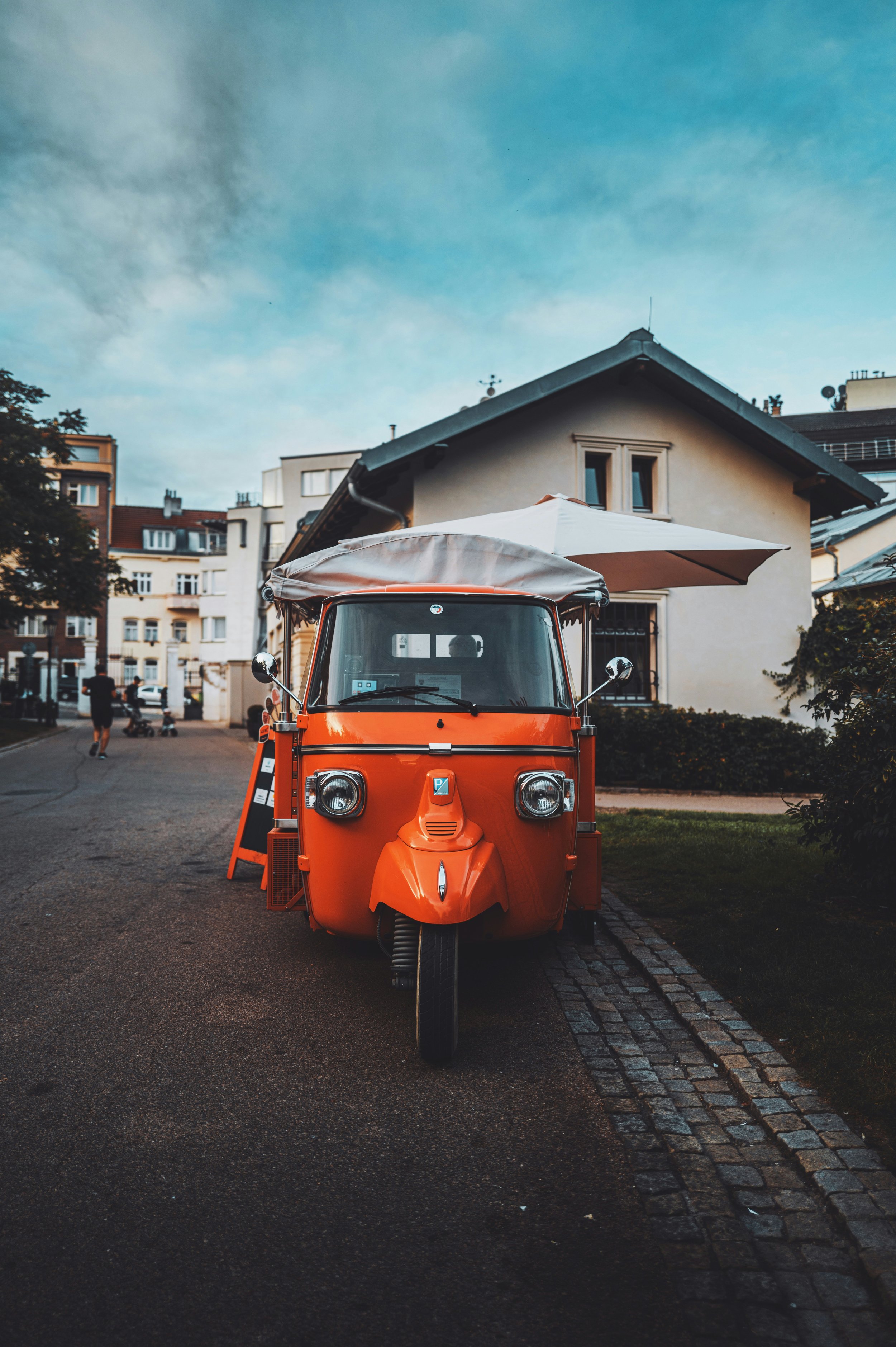Orange three-wheeled food truck with a white umbrella on top, parked on a street in front of residential buildings.