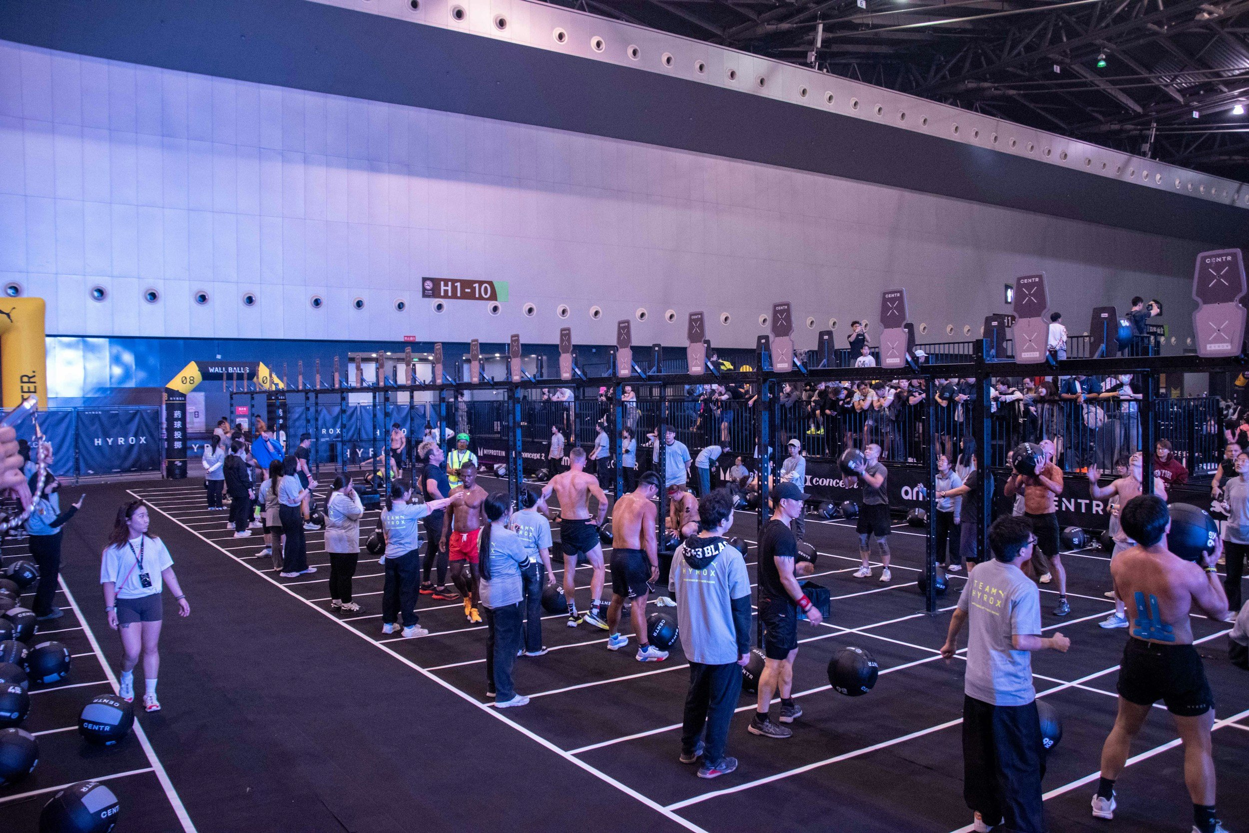 Indoor HYROX fitness event with people exercising, lifting weights, and using combat balls on a large black mat, with workout stations and digital screens in the background.