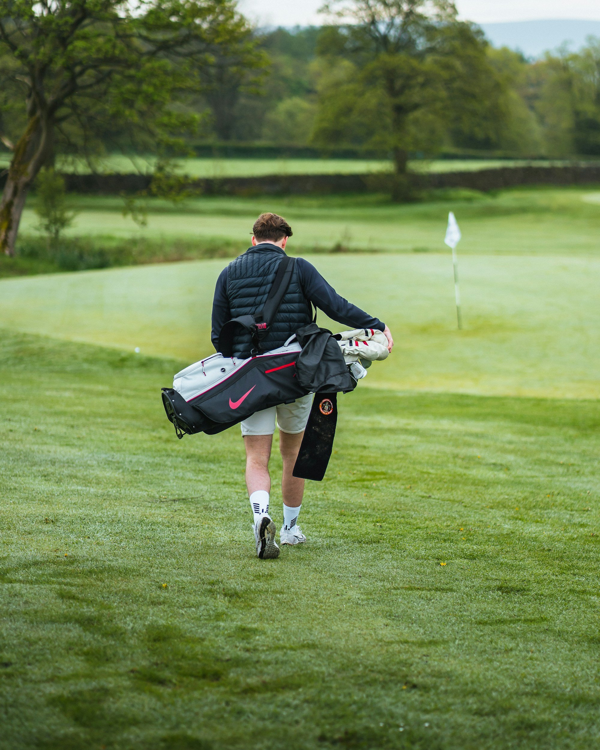 A person walking on a golf course with golf clubs in a bag, wearing a black puffy vest, beige shorts, and white golf shoes, surrounded by green grass, trees, and a white flag on the green.