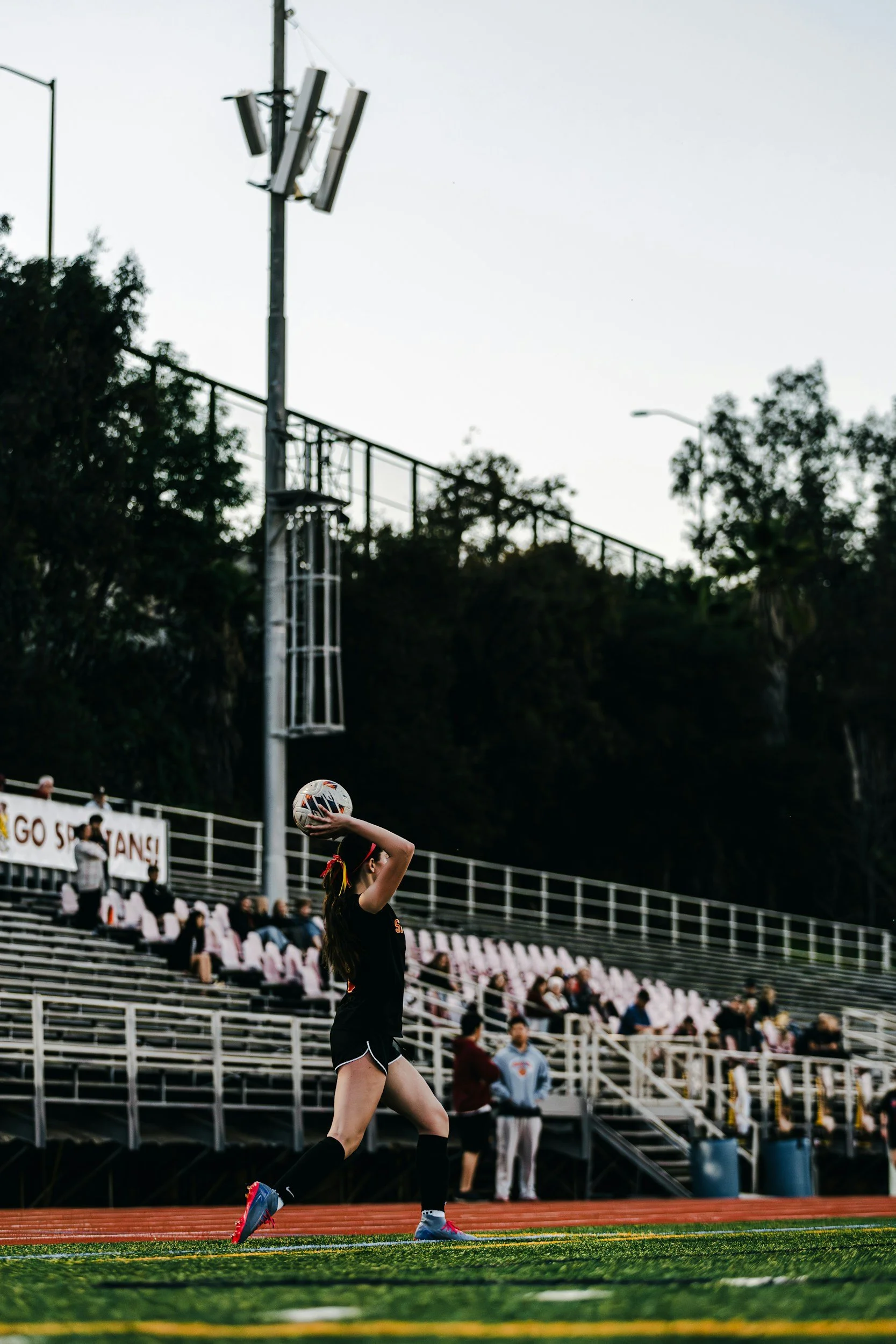 A high school female youth athlete with long hair in a ponytail preparing to kick a soccer ball at a stadium track during a game, with spectators seated in bleachers and a cloudy sky overhead.