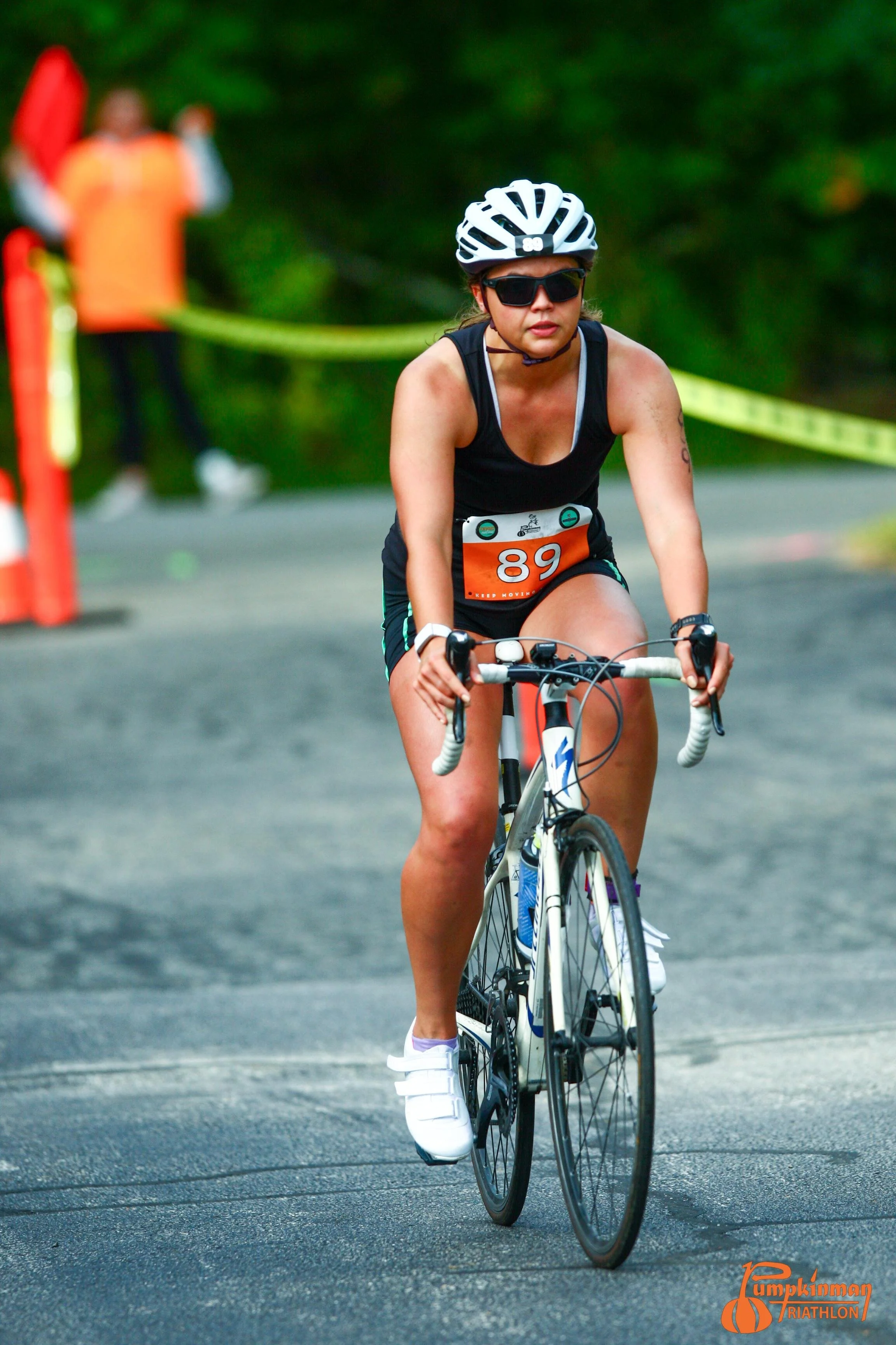 A female cyclist wearing a helmet, sunglasses, and athletic clothing riding a road bike during a triathlon event. There are event markers and a blurred background of trees.