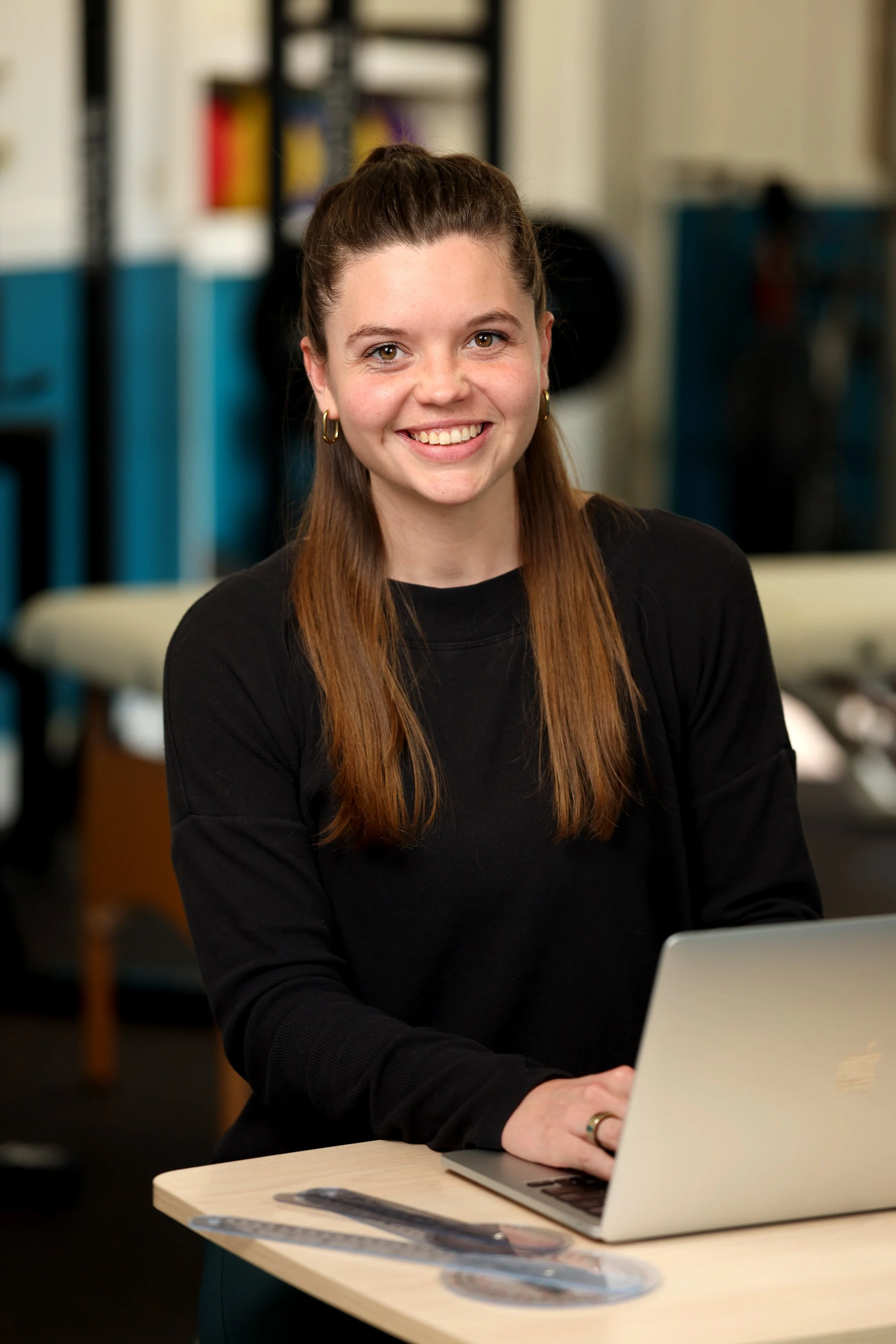 Smiling young woman with brown hair working on a laptop at a standing desk in an physical therapy office setting.
