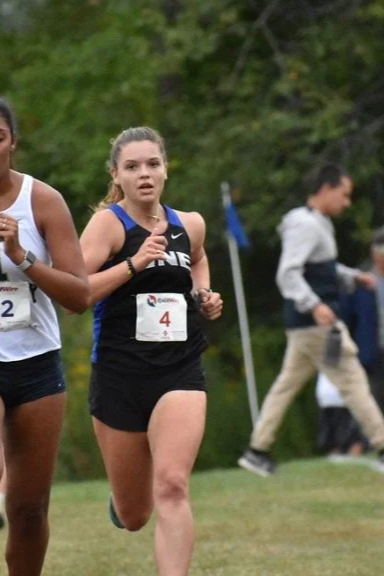 Young female runner in black athletic attire running in a race with other runners and spectators in the background.