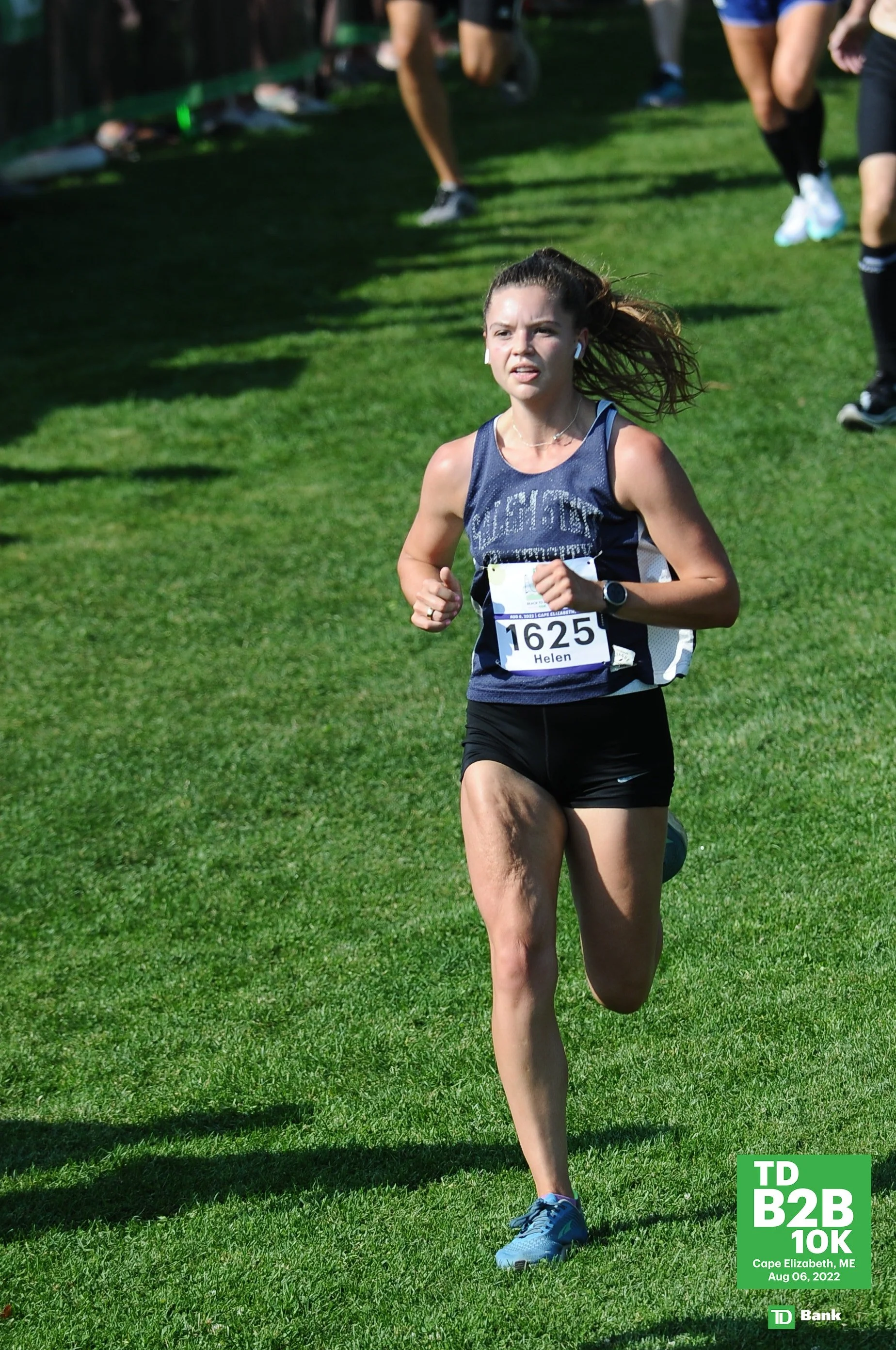 A woman running in a race on a grassy field, wearing a navy blue tank top with the number 1625 and the name Helen, sports earbuds, and blue running shoes. There are other runners and spectators in the background, and a green advertising board in the bottom right corner reads 'TD B2B 10K Cape Elizabeth, ME Aug 06, 2022'.