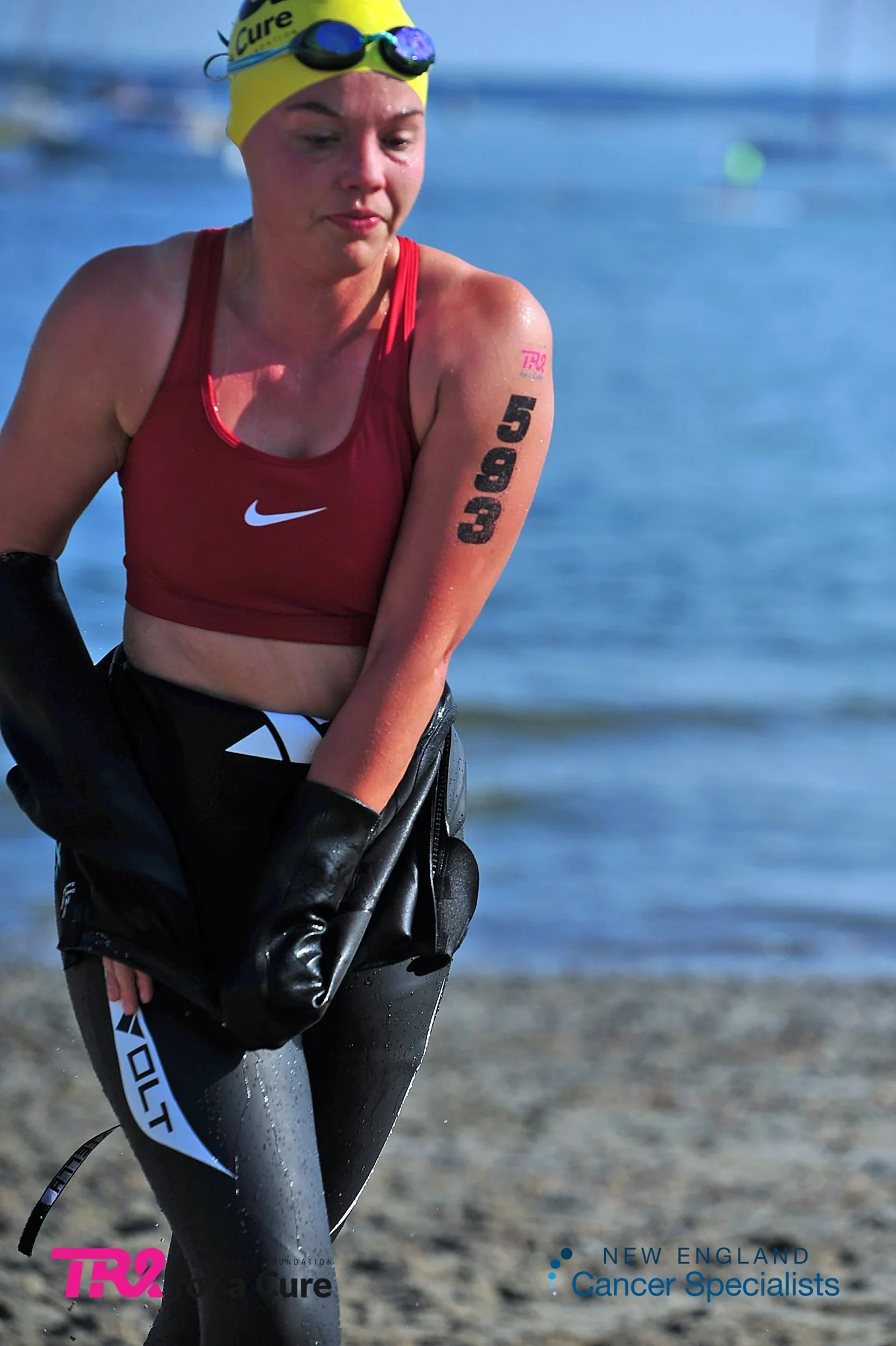 A female athlete on a beach, wearing a yellow swim cap with goggles on top, a red sports bra, and black and gray wetsuit pant. She just completed the open water swim portion of the triathlon Tri for a Cure in Southern Maine