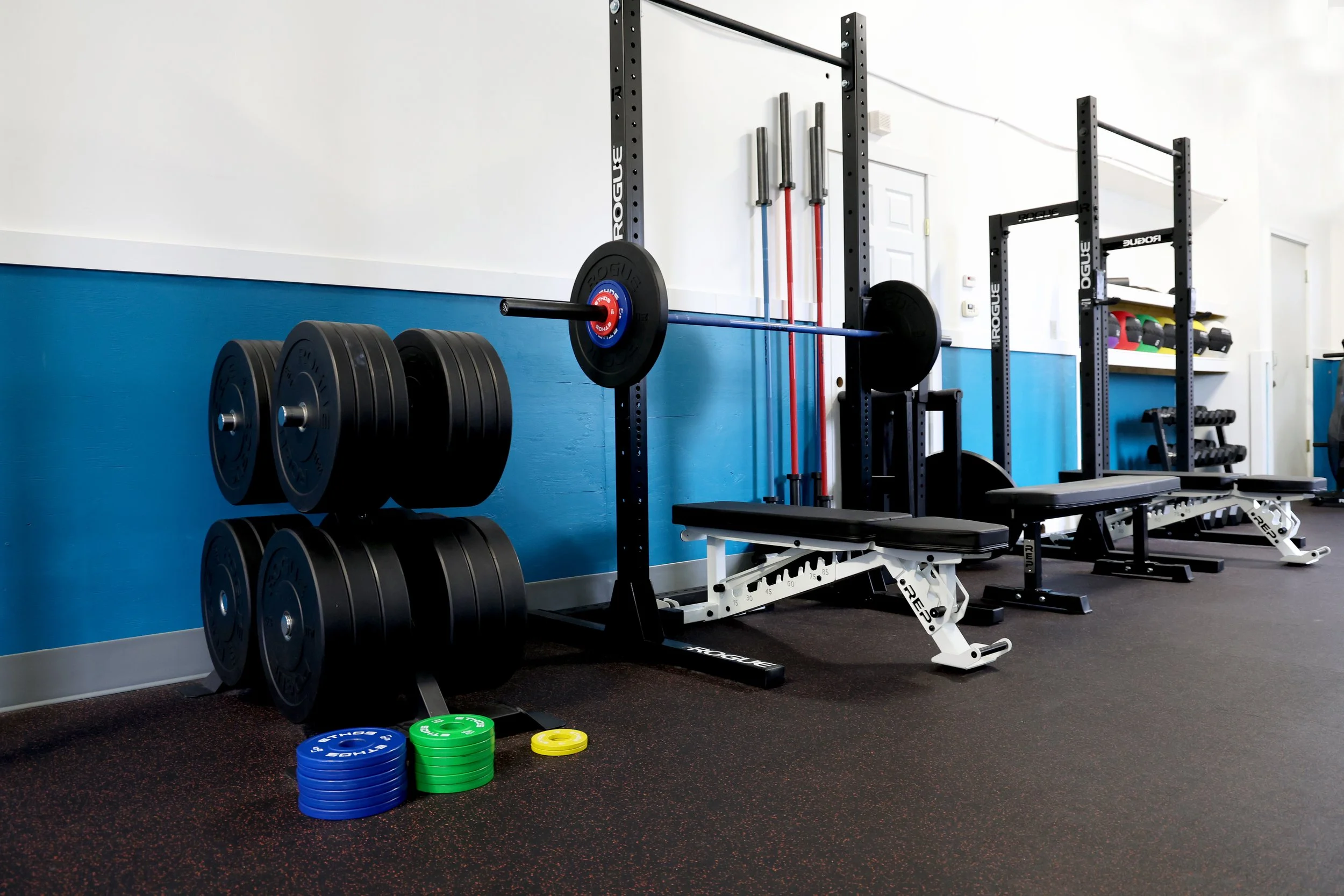 A gym with weightlifting equipment, including barbell with weights, a weightlifting bench, and various colored weight plates on the floor.