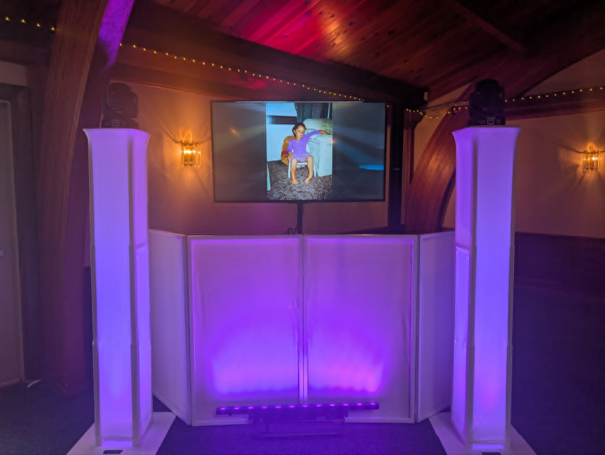 A large indoor space with purple-lit columns, a wooden arched ceiling, a large screen displaying a woman in a purple dress sitting on furniture, and decorative string lights on the ceiling.