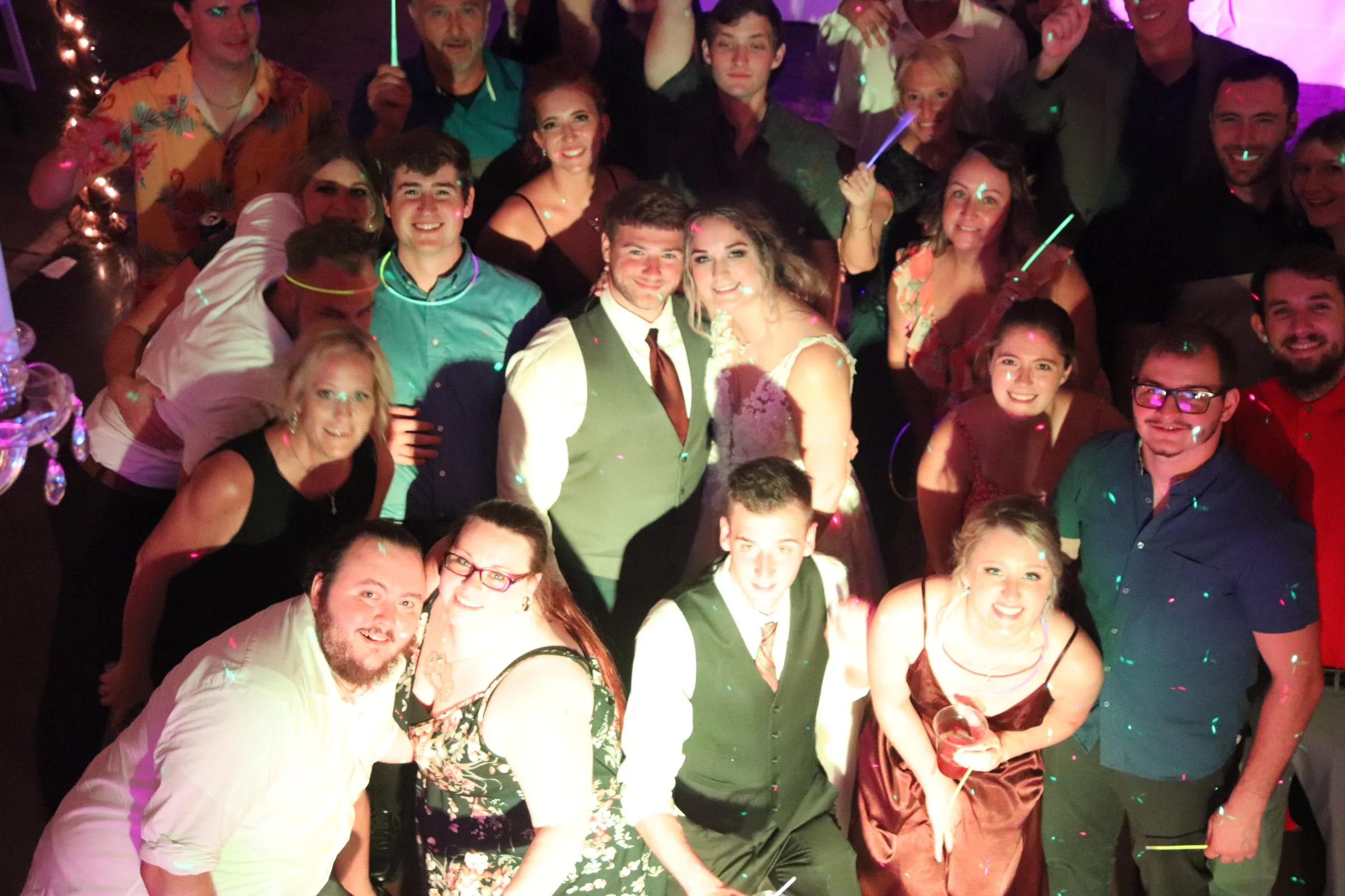 Group of people at a party or celebration, smiling, with some holding drinks. The environment has colorful lights and a festive atmosphere.