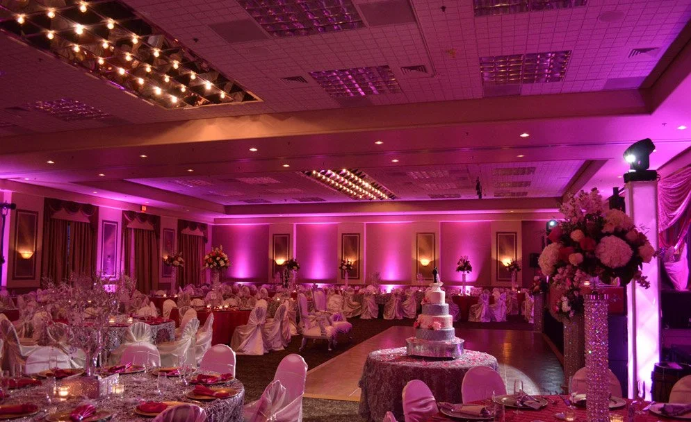 An elegantly decorated banquet hall with pink lighting, round tables covered with pink cloths, floral centerpieces, and a multi-tiered wedding cake.