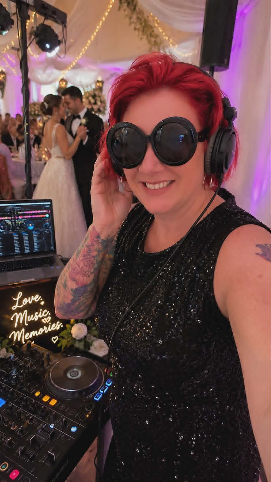 A woman with red hair and tattoos smiling while wearing DJ headphones and sunglasses at a wedding reception. In the background, a bride and groom dance, decorated with fairy lights and floral arrangements.