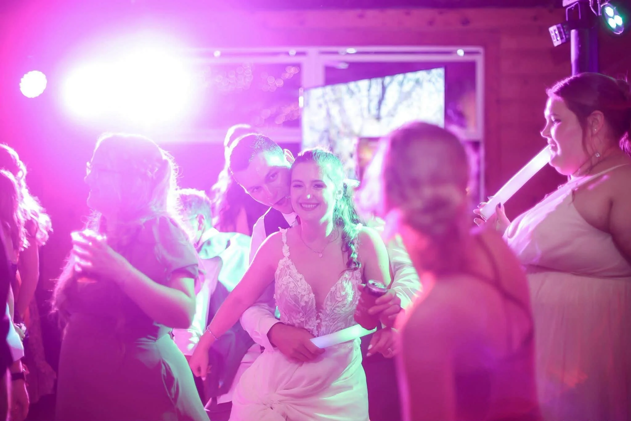 A wedding celebration scene featuring a wedding cake decorated with pink and white designs, surrounded by an abundance of pink, purple, and cream flowers, with two glasses of wine and a small calendar on the table, against a scenery wallpaper depicting countryside houses and trees.
