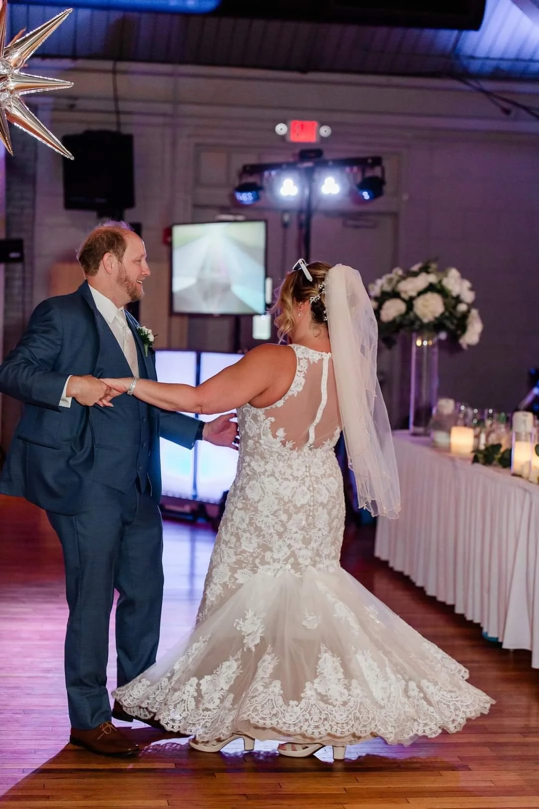 A bride and groom dancing at their wedding reception in a decorated venue with candles and floral arrangements.