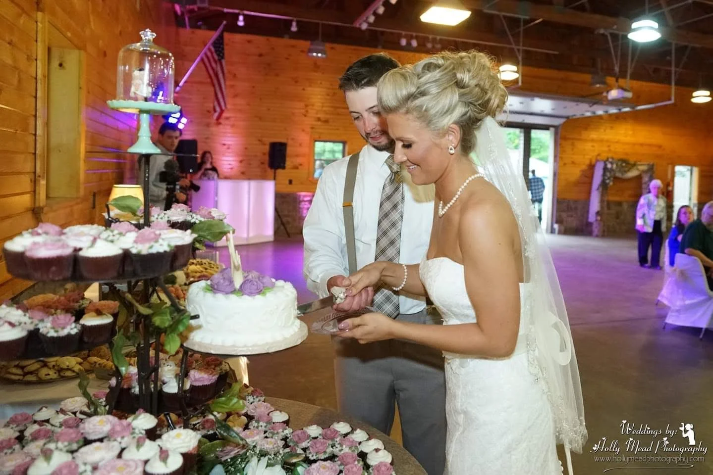 Bride and groom cutting their wedding cake at a reception with desserts on a table in front of them, wooden walls, and other guests in the background.