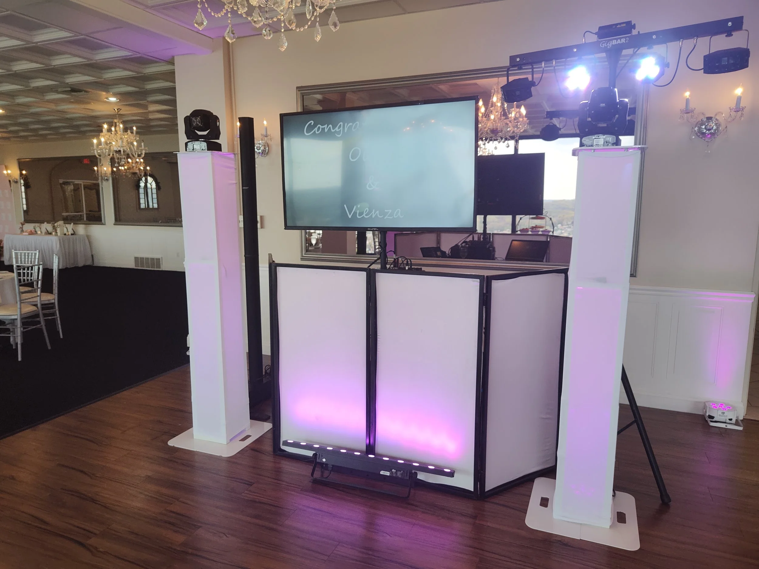 Wedding reception setup with a dance floor, white pillars with purple lights, a large screen displaying congratulatory message, DJ equipment, and chandeliers.