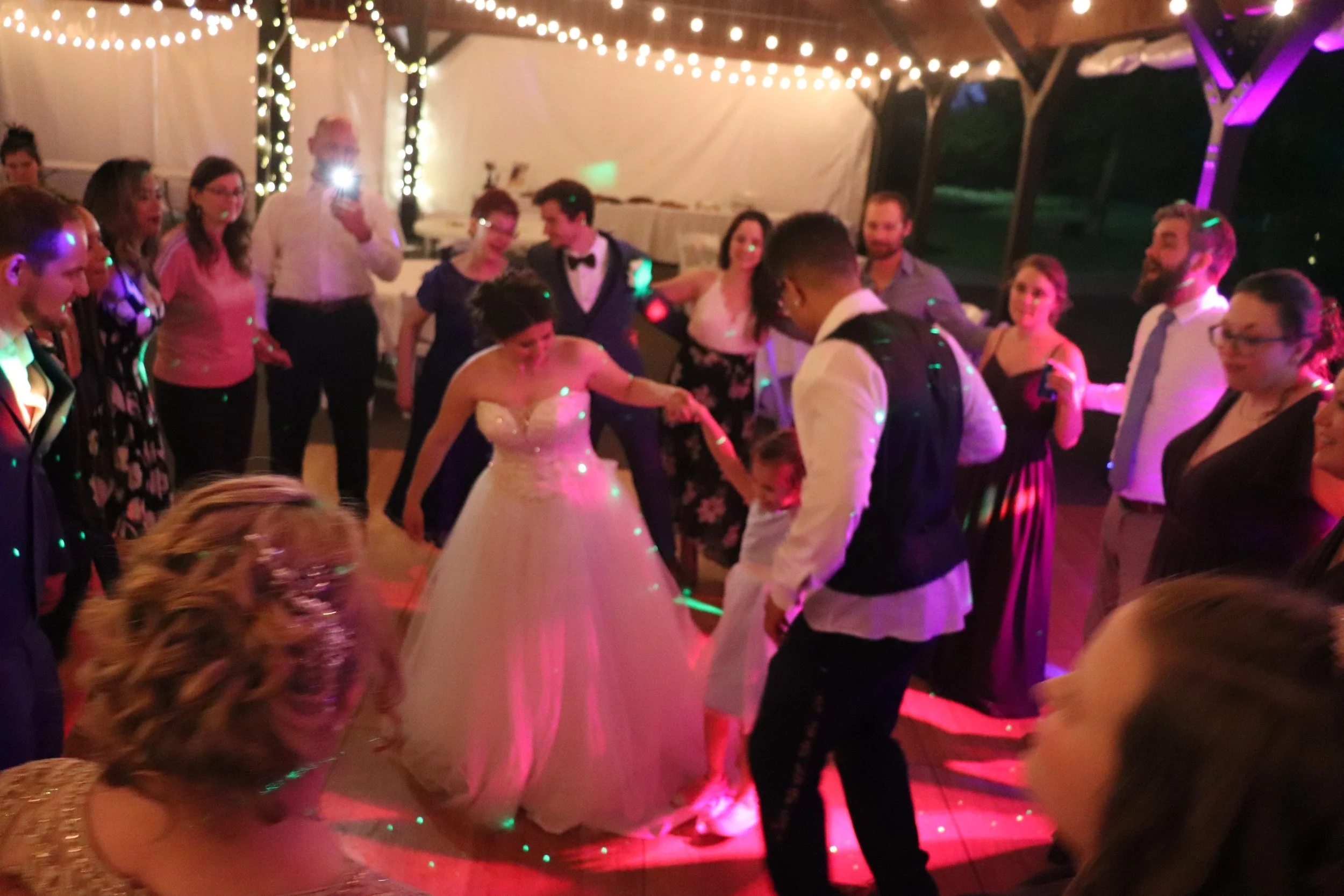 A group of people dancing and celebrating at an indoor wedding reception under string lights. The bride in a white gown and the groom in a vest are at the center of the dance floor, surrounded by friends and family.