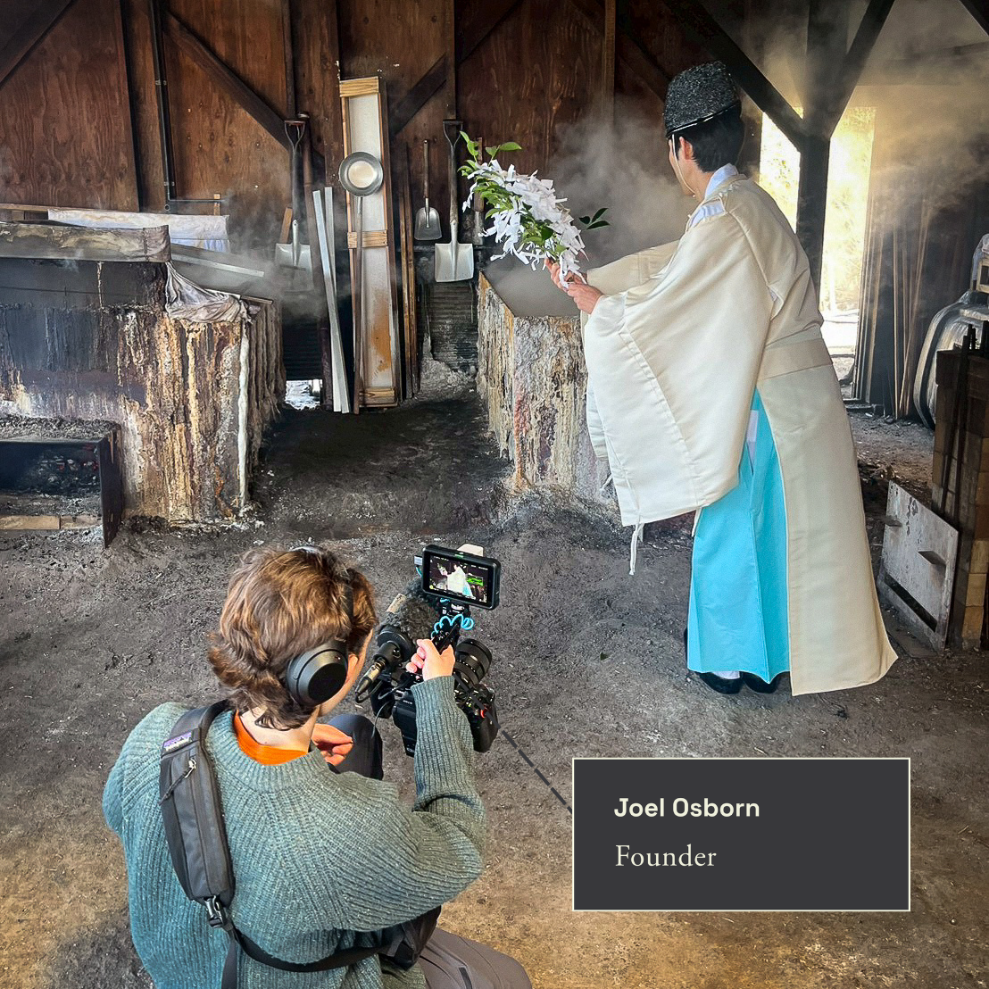 A person dressed in traditional Japanese attire performing a ritual in a rustic setting with a camera operator filming the scene. The performer stands beside a stone altar or fire pit, holding a branch with flowers, with a cloud of smoke or steam rising behind them.