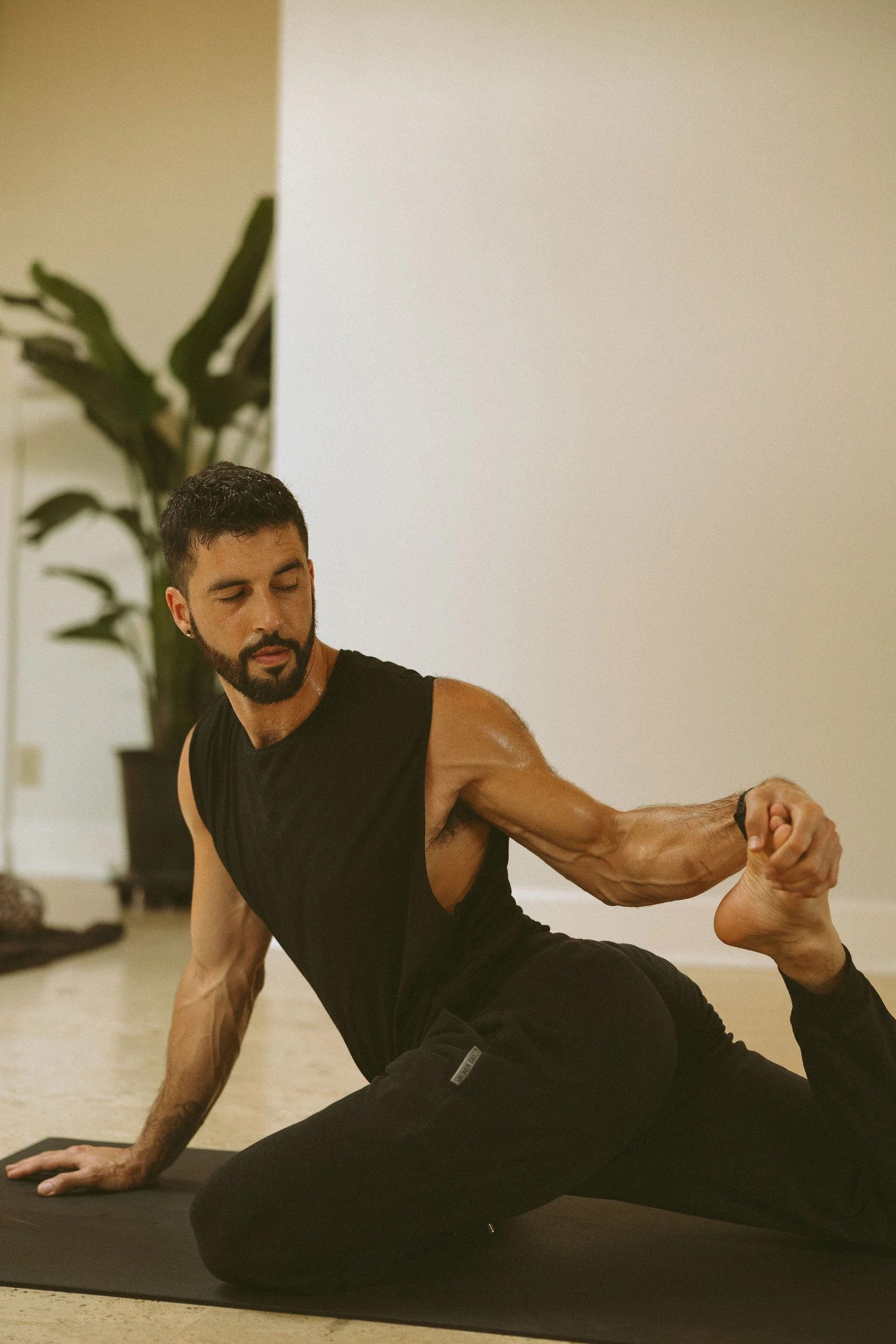 A man with a beard and short dark hair practicing a yoga pose indoors. He is on a yoga mat, wearing a black sleeveless shirt and black pants, with a large plant in the background.