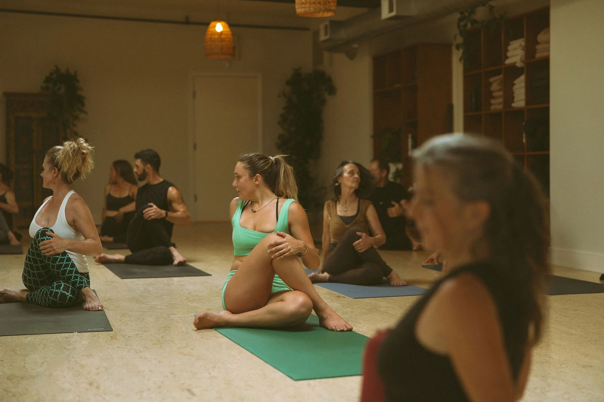 Group of people practicing yoga on mats in a studio with wood shelves and plants, some individuals in the foreground and background, engaging in stretches and poses.