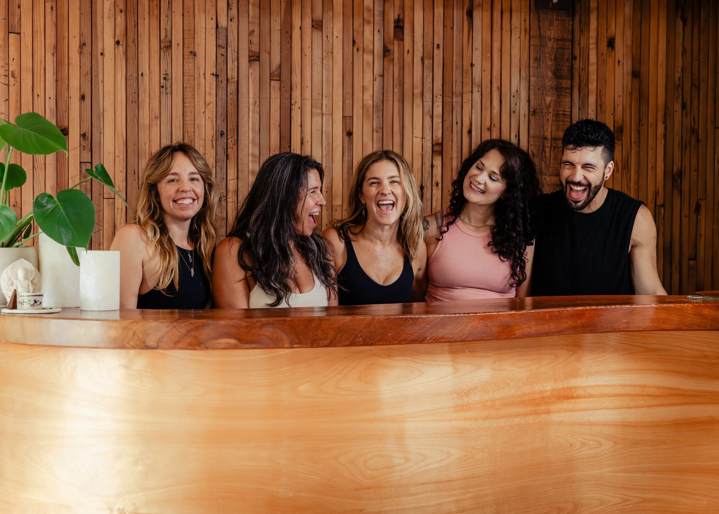 A group of five people, four women and one man, sitting and laughing together in front of a wooden wall, with a large green plant and some white decorative items on a table in the foreground.