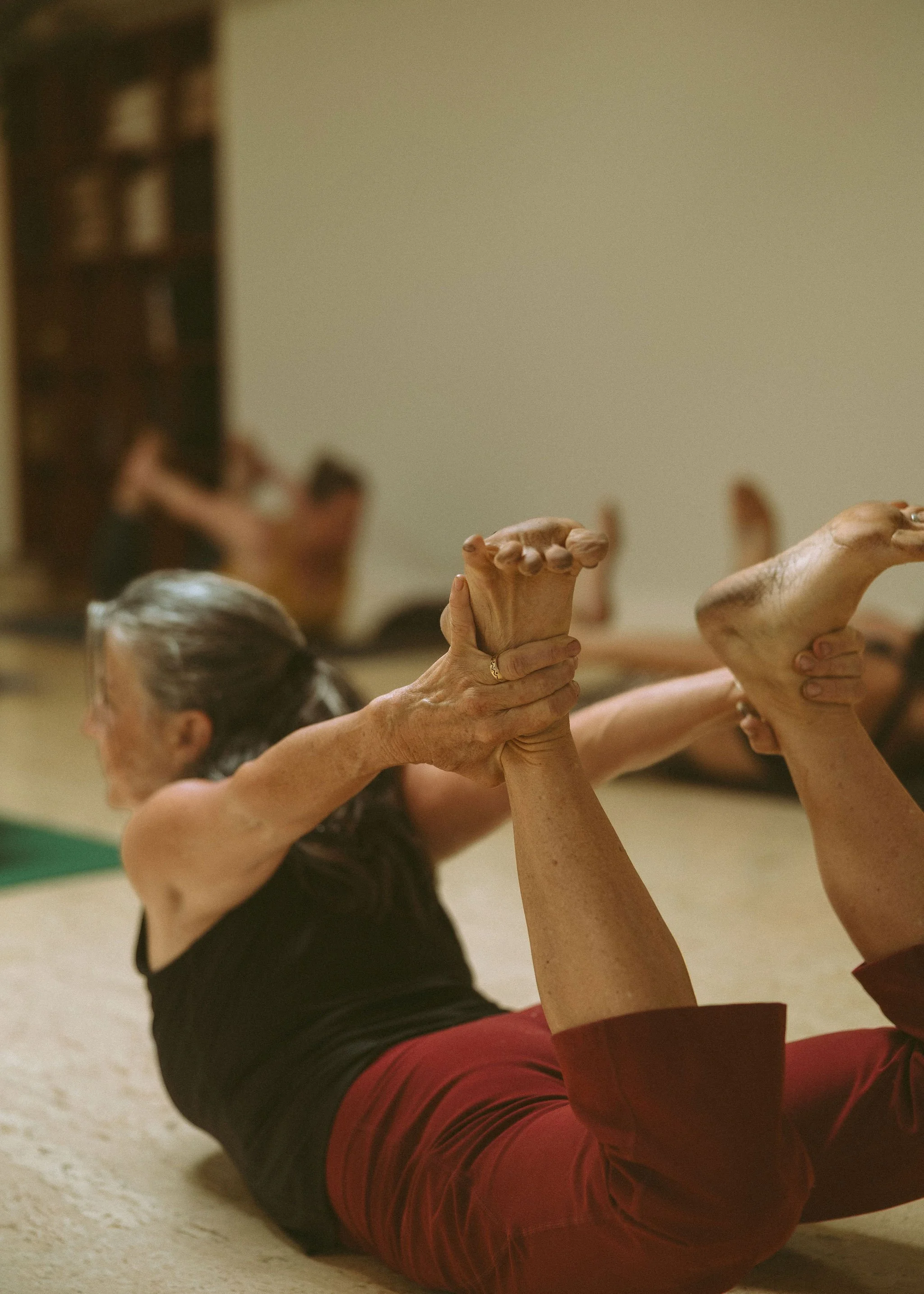 Older woman in a black tank top and red pants practicing yoga, balancing on her back with her legs raised and her hands clasped together, in a room with a plain wall and hardwood floor.