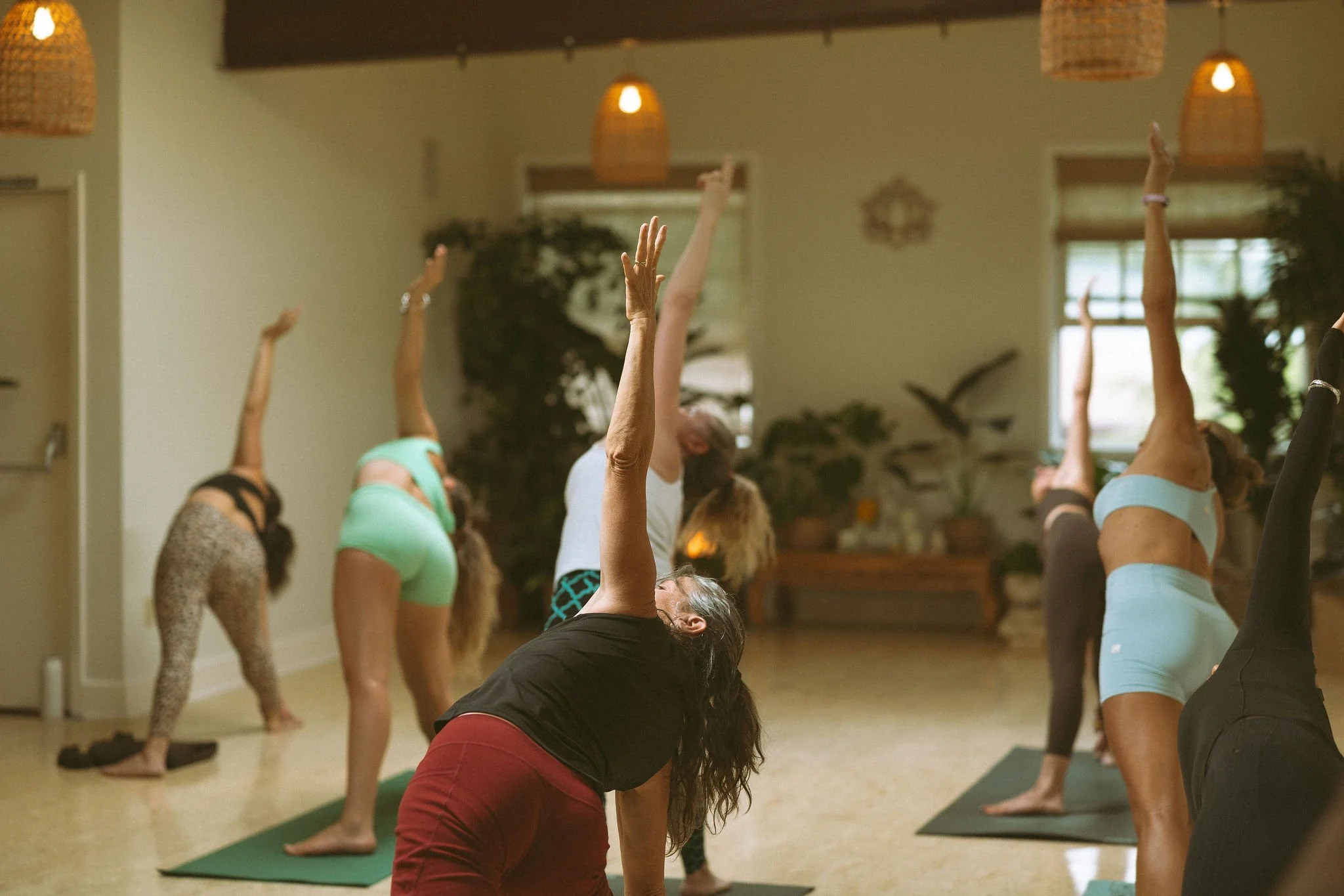 Group of people practicing yoga in a studio, some in downward dog pose and others stretching their arms, with natural light coming through windows.