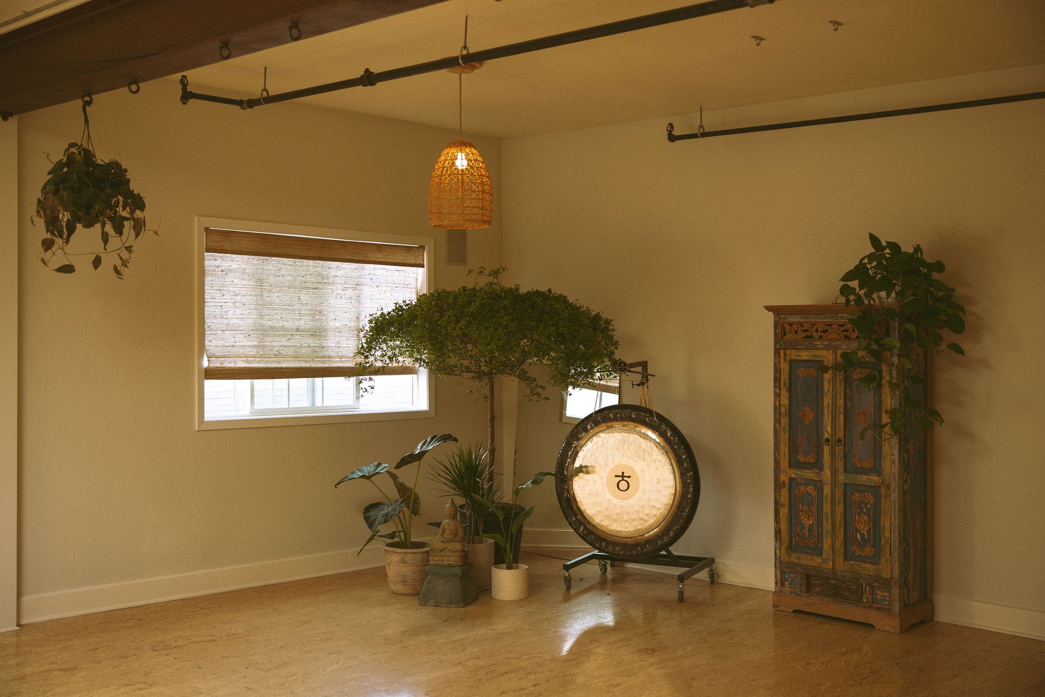Interior of a room with large windows, potted plants, a decorative cabinet, and a hanging wicker lampshade.