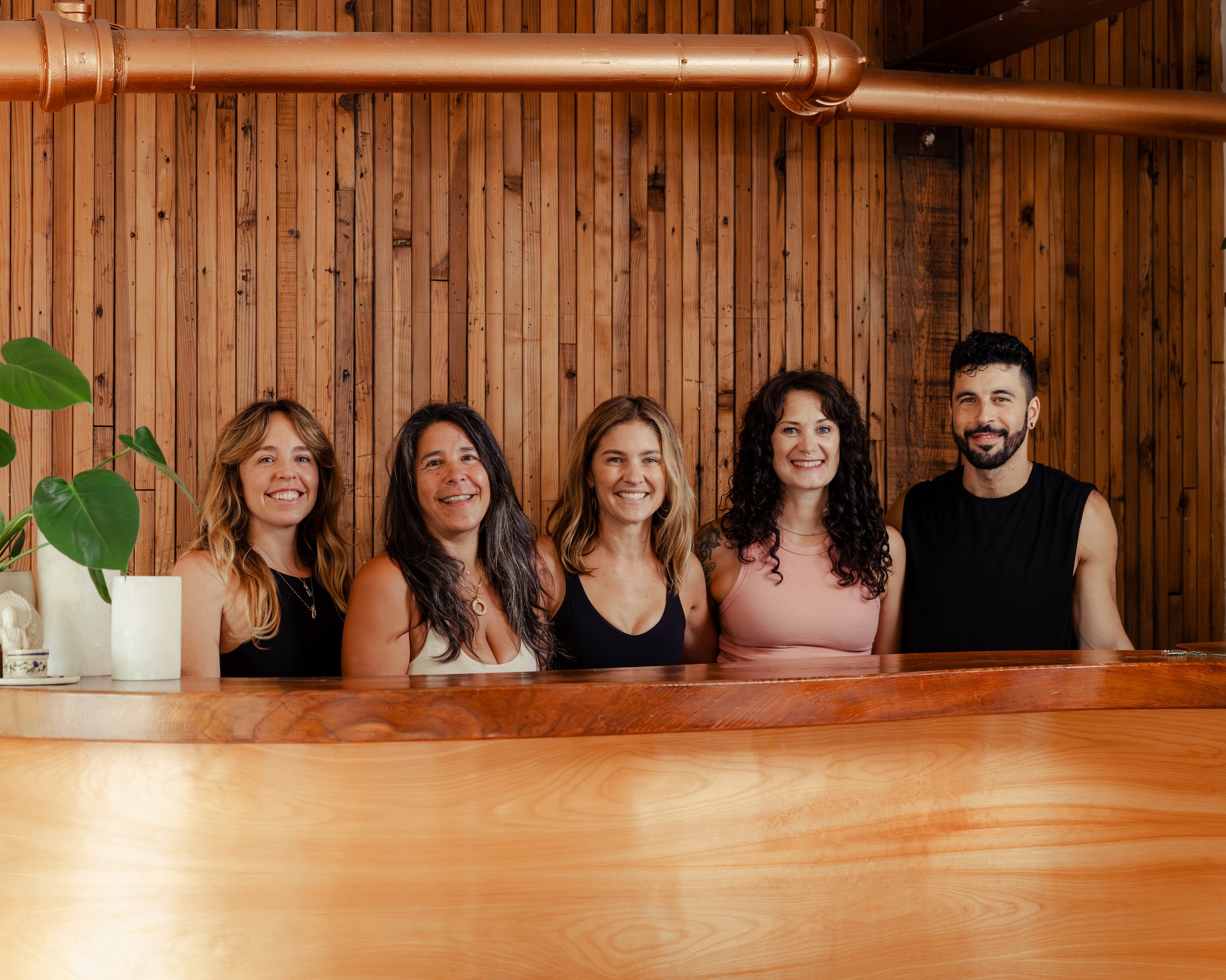 Group of five women and one man sitting at a wooden table, smiling, with a wooden wall and a plant in the foreground.