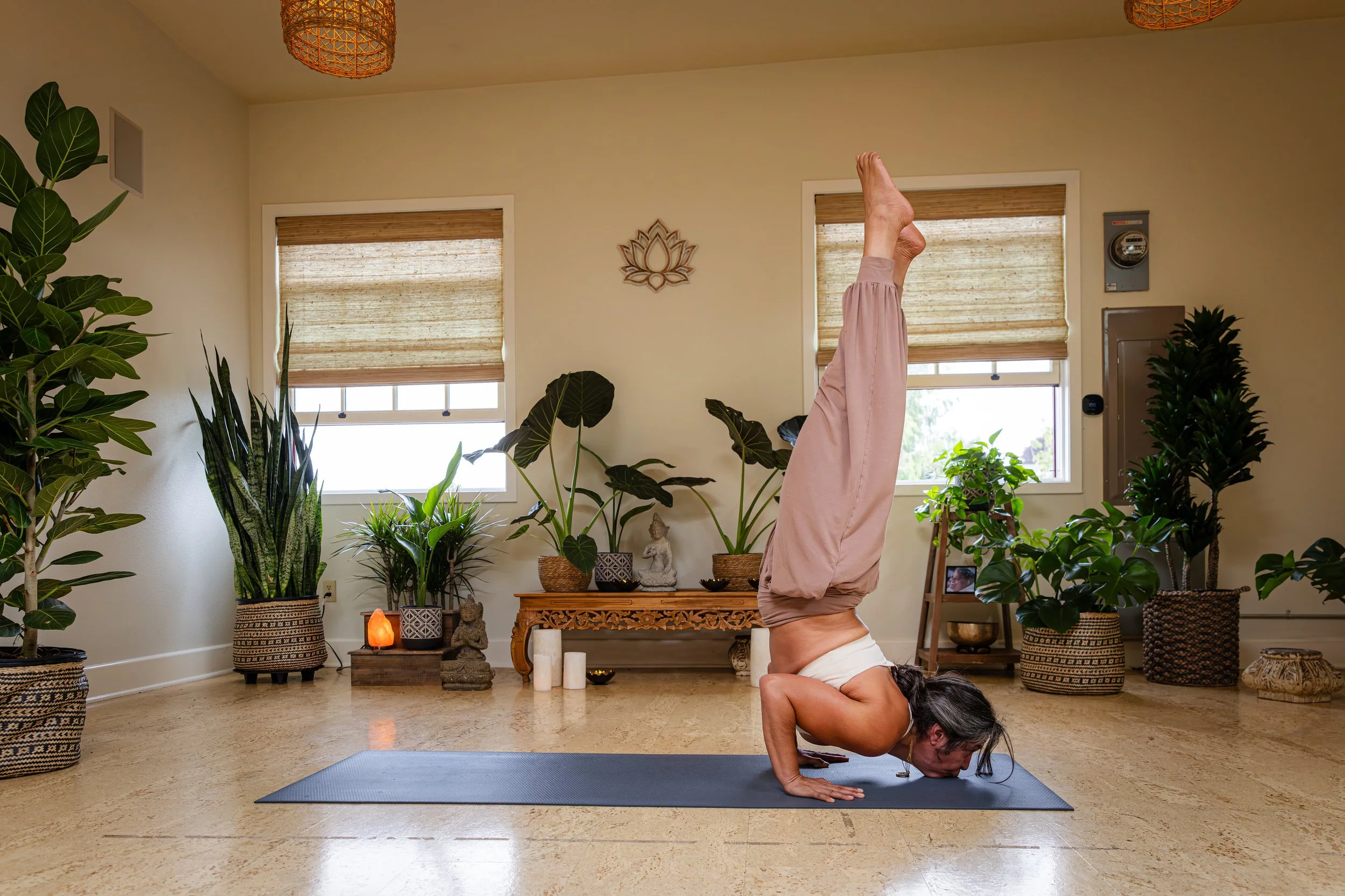 A woman is doing a headstand yoga pose in a room filled with plants and decorative items, with her hands on the floor and legs extended upwards. She is on a yoga mat on a wooden floor.