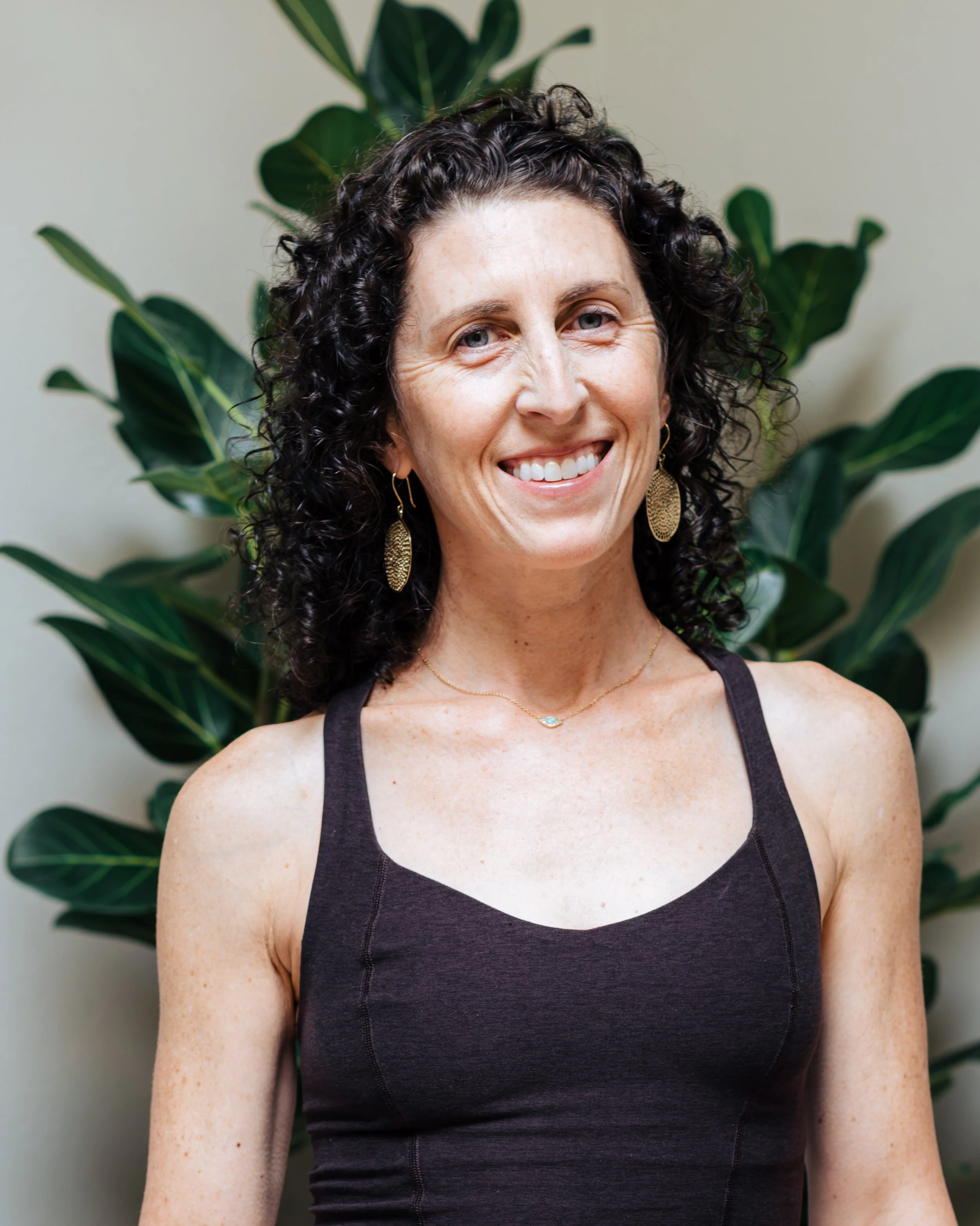Close-up of a smiling woman with curly dark hair, wearing a black sleeveless top, gold earrings, and a delicate necklace, standing in front of large green leafy plants.