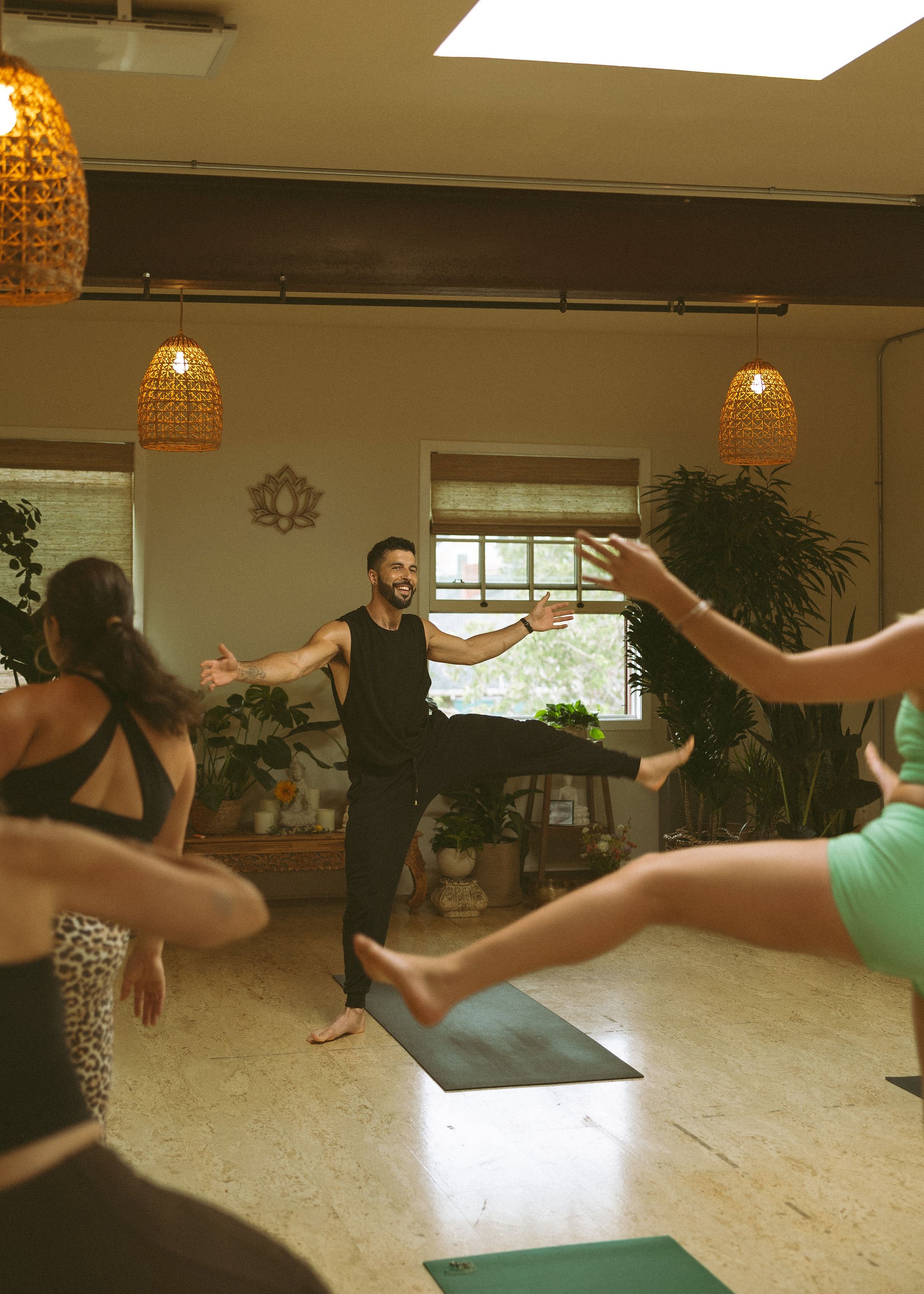 A group of people participating in a yoga or dance class in a studio with natural lighting, plants, and decorative hanging lights. A male instructor is demonstrating a pose with arms outstretched and one leg lifted.