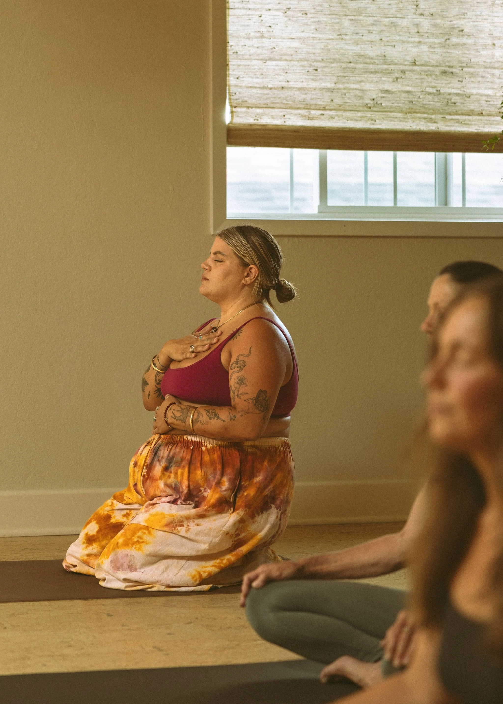 A woman with tattoos on her arms and shoulder, wearing a pink tank top and colorful tie-dye pants, sitting on the floor with eyes closed and hand on her chest, engaging in a meditation or yoga session in a room with natural light coming through a win